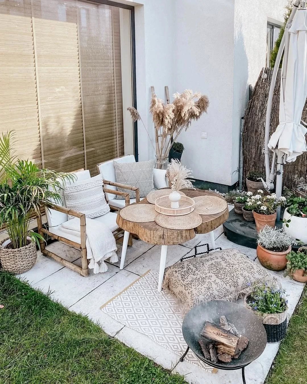 Outdoor patio with wooden chairs, a round table, cushions, plants, potted flowers, and a fire pit offers charming patio decoration on a tiled surface next to a white building with neutral decor.