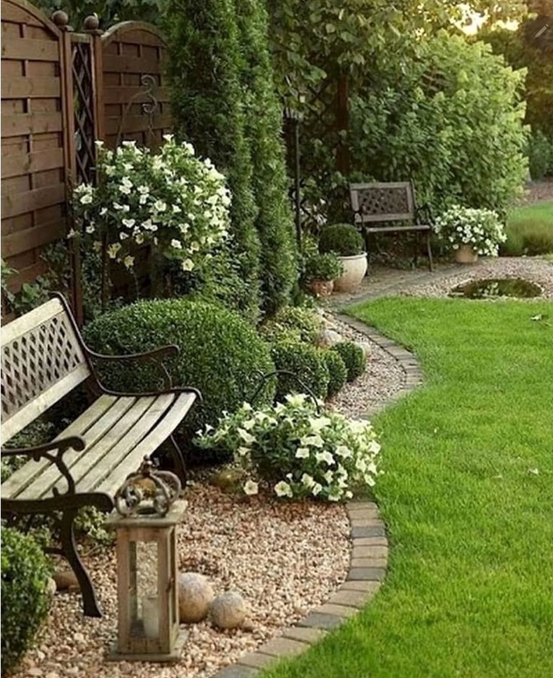 A landscaped garden with benches, gravel paths, potted white flowers, trimmed bushes, and green grass bordered by a wooden fence and hedges offers inspiring small backyard landscaping for any home.