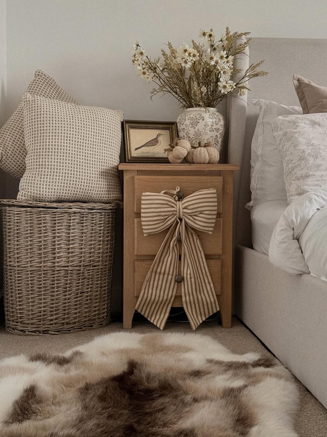 A wooden nightstand with a striped bow, vase of flowers, framed bird print, pumpkins, and pillows sits beside a bed with a fur rug—perfect for adding vintage farmhouse decor charm to your space.
