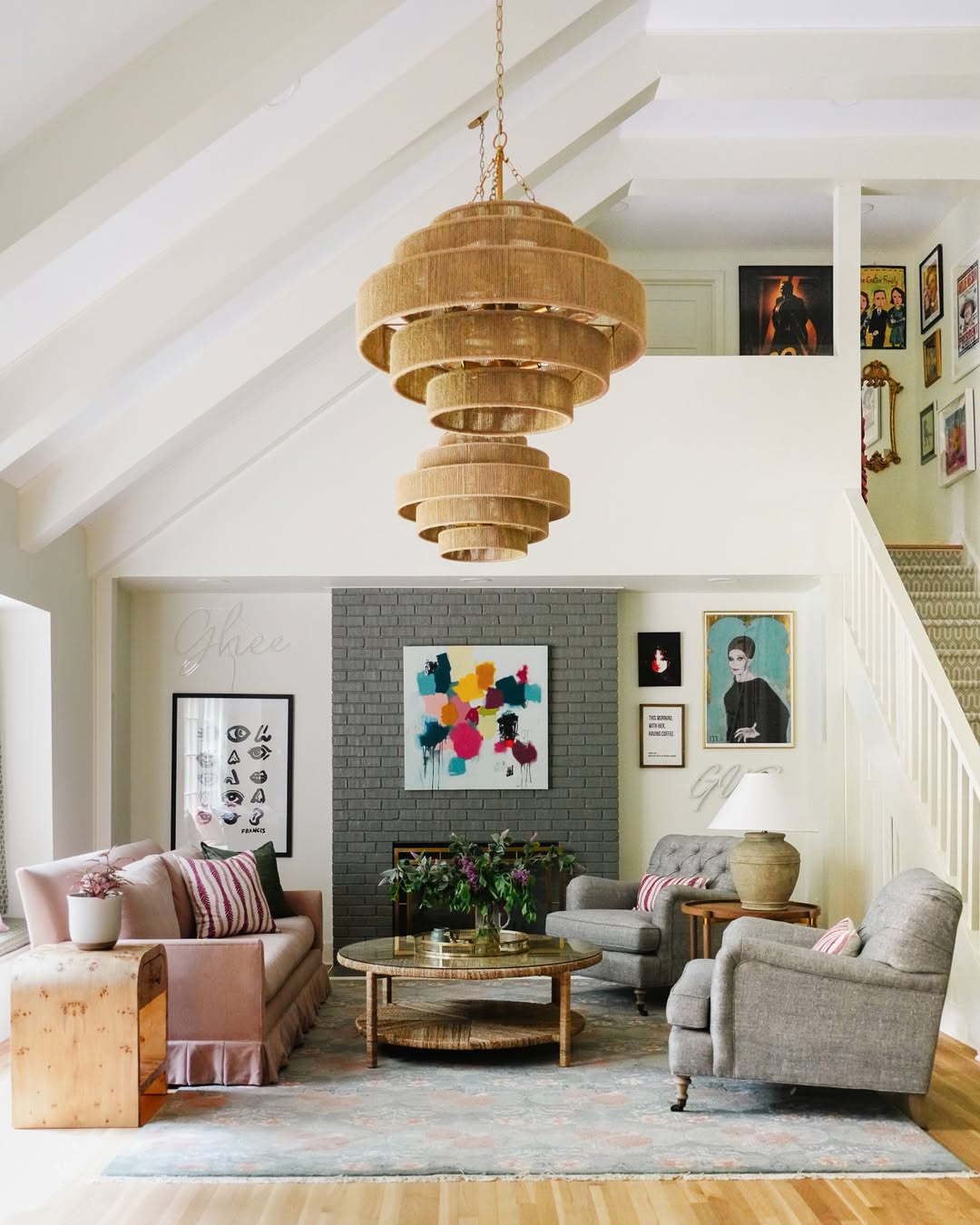 A stylish modern living room features a beige sofa, gray armchairs, a circular coffee table, colorful artwork, and a large layered chandelier under a white vaulted ceiling.