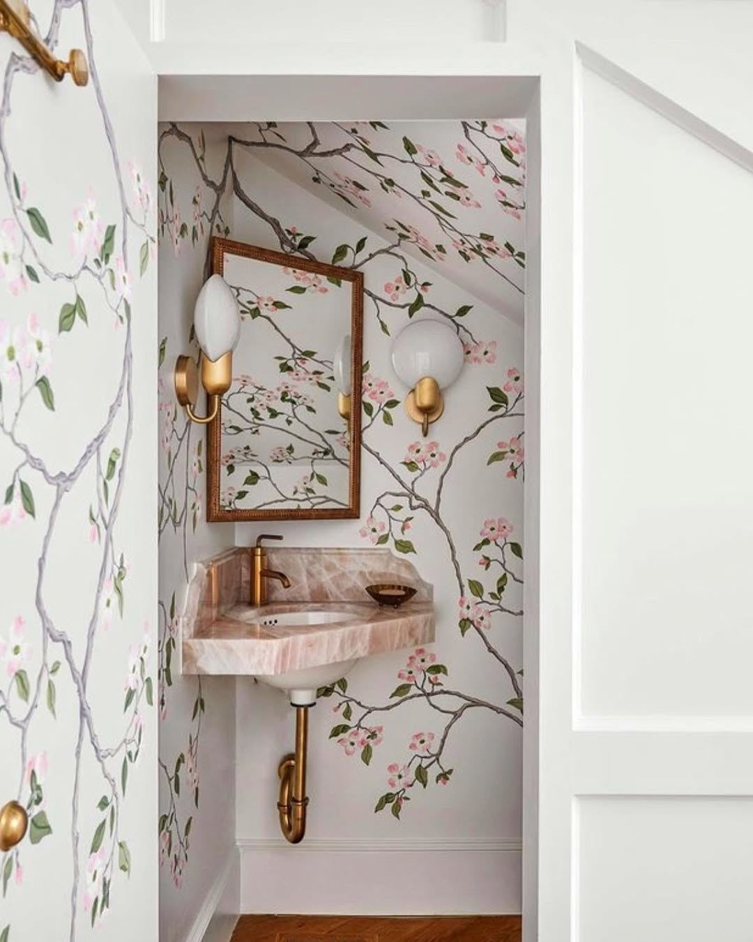 This stylish powder room features floral wallpaper, a pink marble corner sink with gold fixtures, a rectangular mirror, and wall sconces under a sloped ceiling—perfect for wow-factor inspiration and chic powder room ideas.