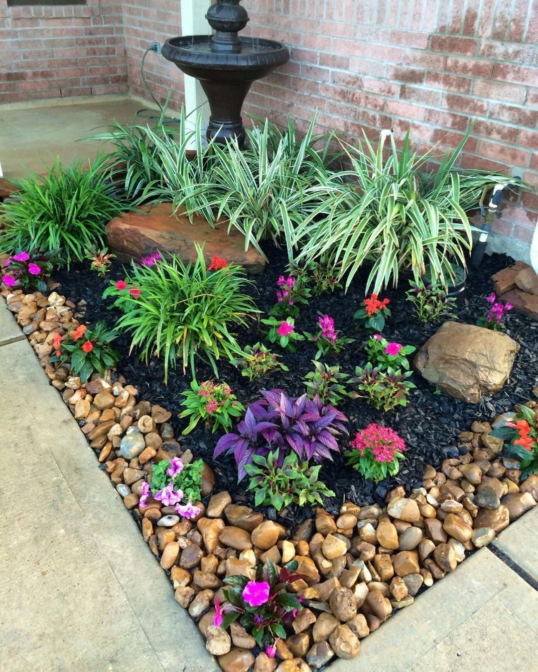 A small garden bed with green and purple plants, colorful flowers, rocks, black mulch, and a water fountain against a brick wall offers inspiring small backyard landscaping ideas.
