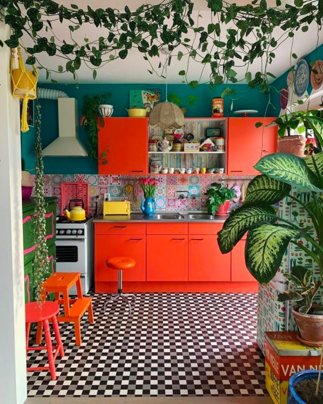 Colorful orange kitchen with red cabinets, patterned backsplash, black and white checkered floor, orange stools, and various potted plants hanging and placed around the room.