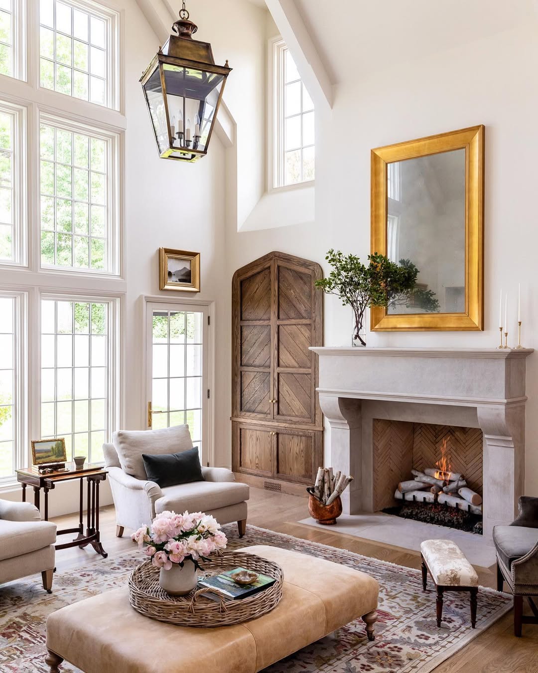 Bright high ceiling living room with tall windows, light armchairs, a fireplace with a gold-framed mirror, wooden armoire, and a large ottoman topped with flowers and books—perfect inspiration for bigger home living.