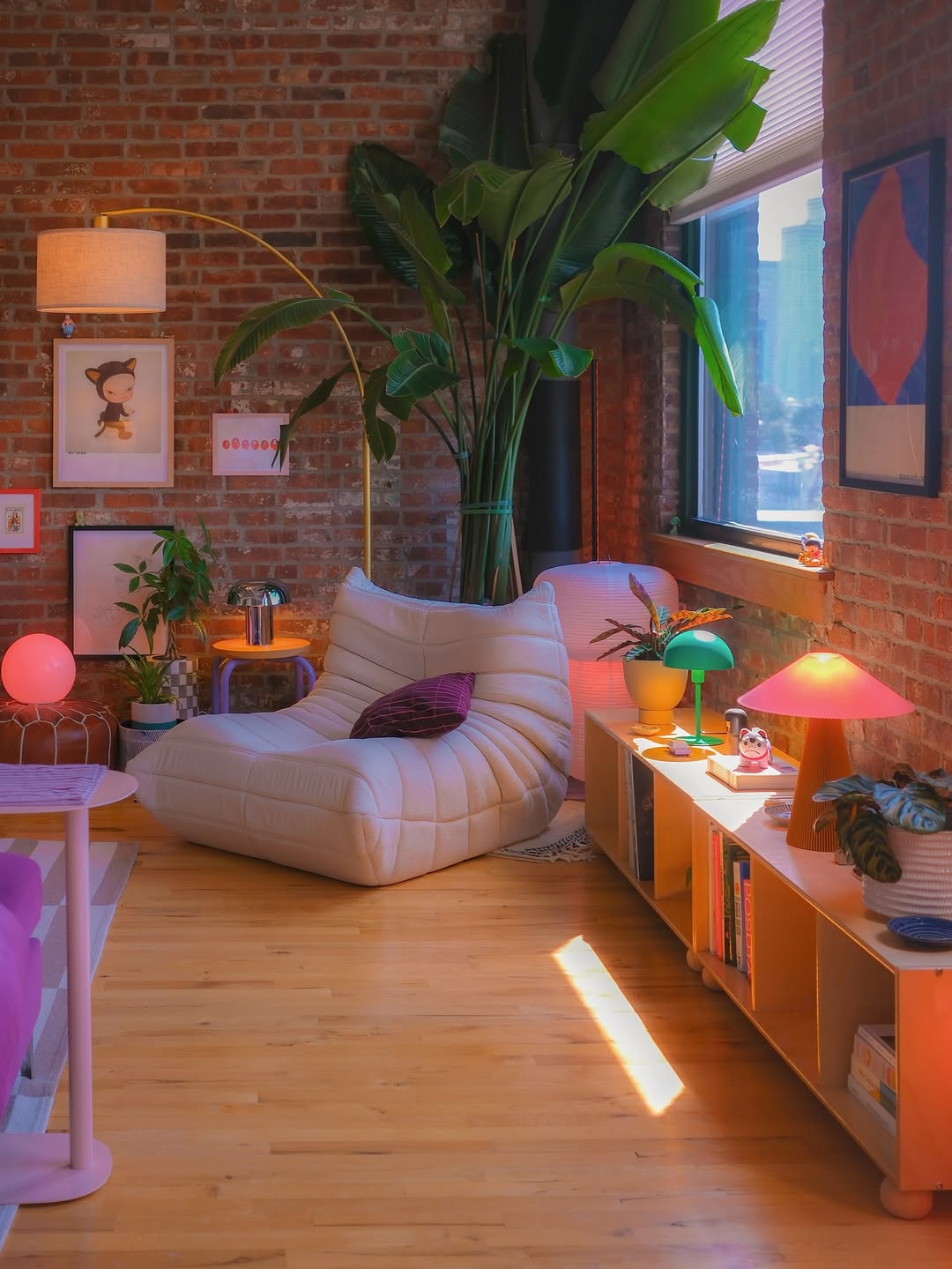 A cozy modern living room with exposed brick walls, a large leafy plant, a white lounge chair, colorful lamps, and a low bookshelf by a sunlit window—perfect inspiration for your own 30 modern living room ideas.