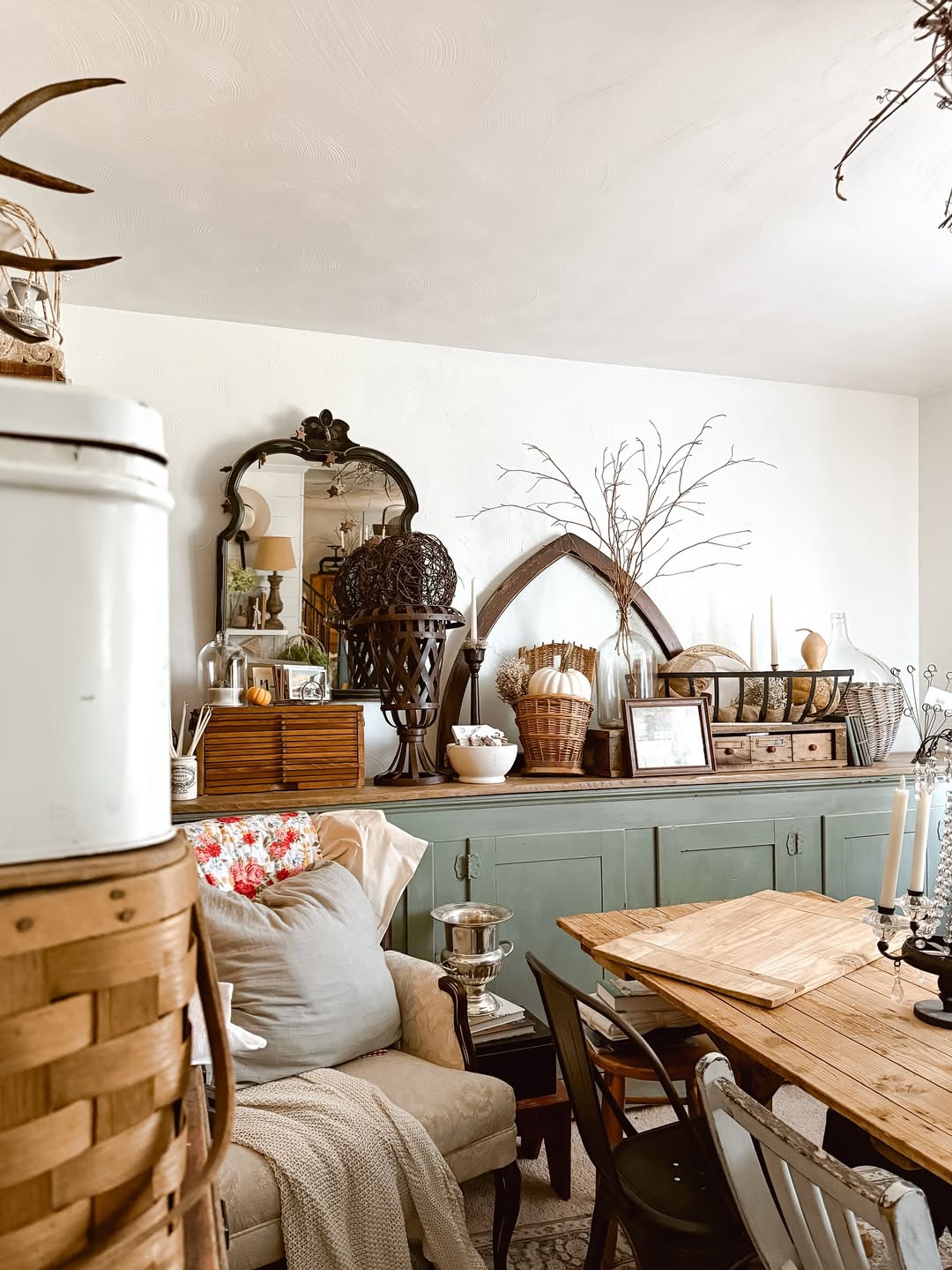 A cozy dining and living area featuring a wooden table, mixed chairs, a cushioned armchair, green sideboard, mirrors, baskets, candlesticks, and rustic accents—perfect inspiration for vintage farmhouse decor lovers.