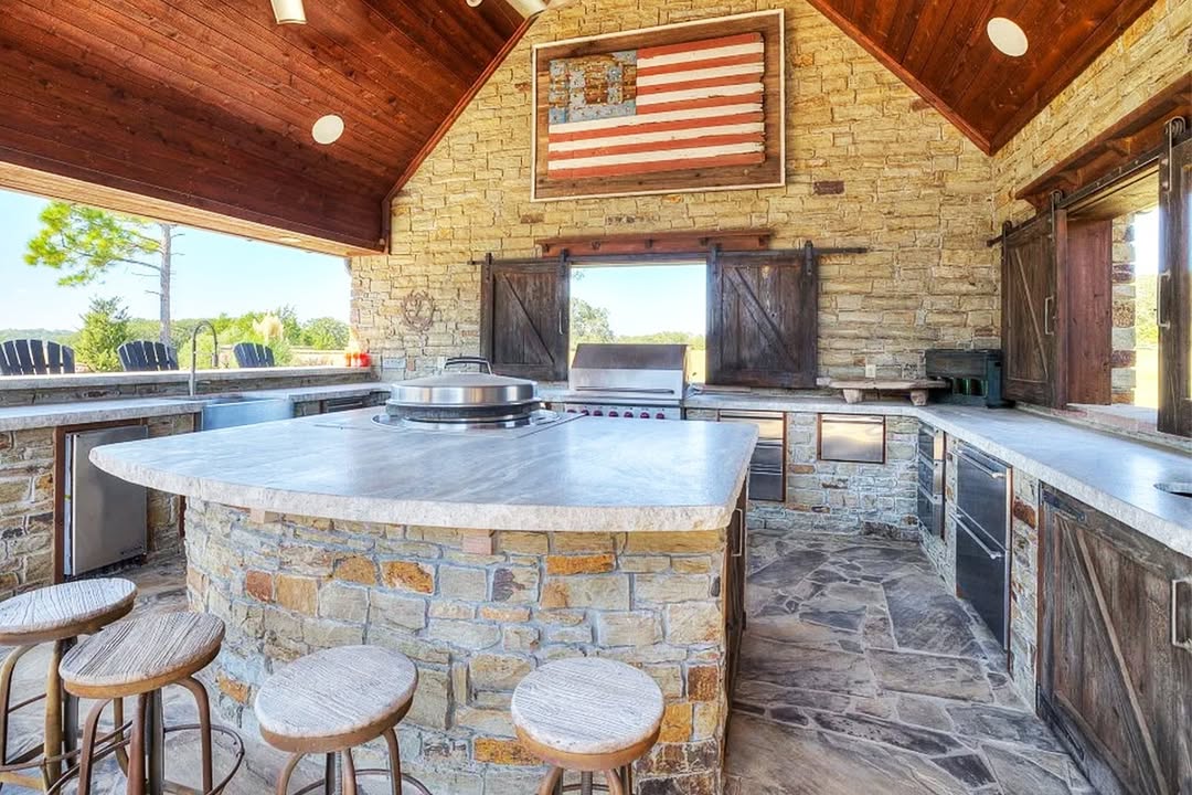 Rustic outdoor kitchen with stone walls, marble countertops, bar stools, stainless steel appliances, and a large American flag artwork on the back wall—perfect for those seeking inspiring outdoor kitchen ideas.