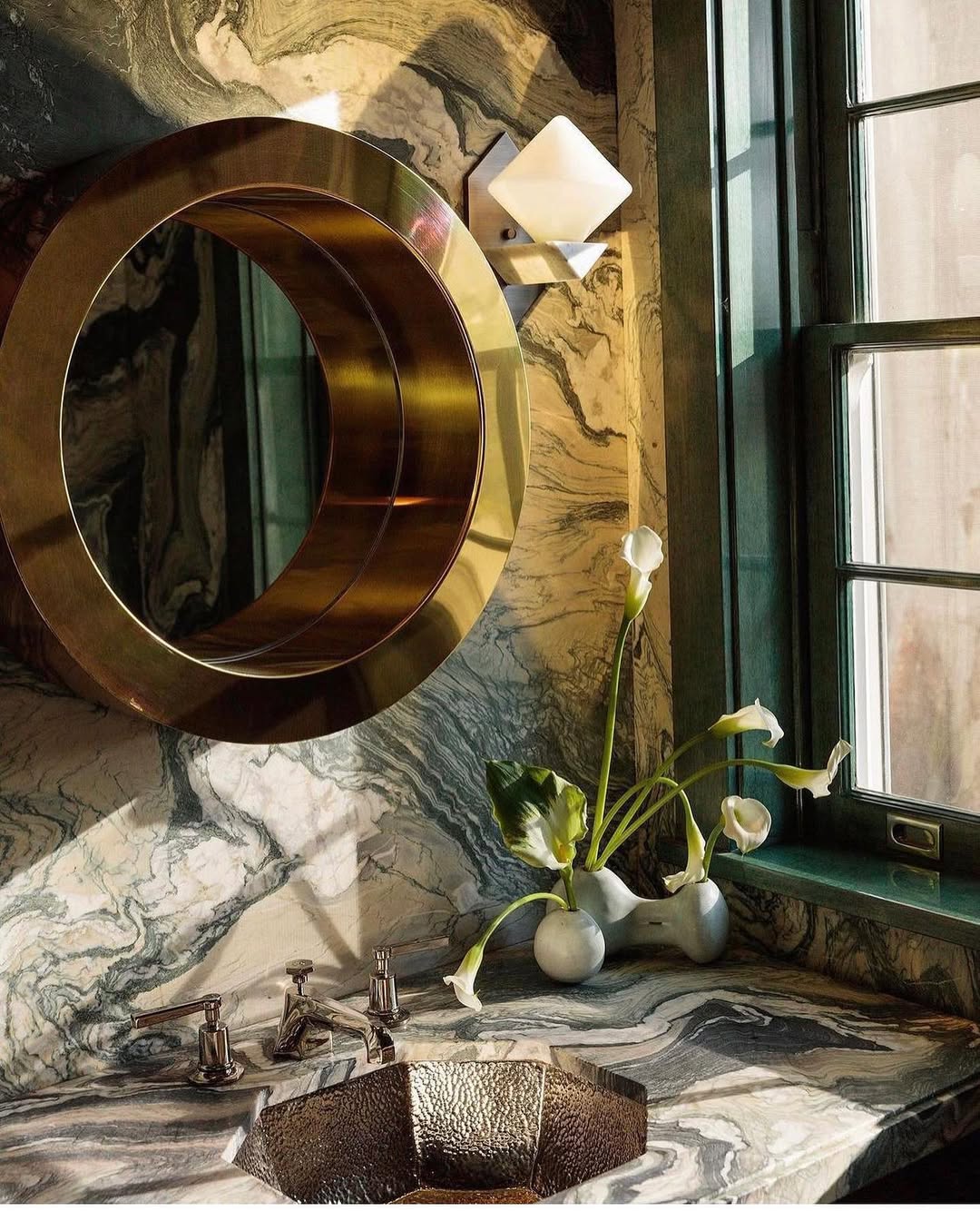 Modern bathroom with a round brass mirror, marble countertop and walls, metallic sink, wall sconce, and a vase with white flowers near a window—perfect inspiration for stylish powder rooms with wow-factor.