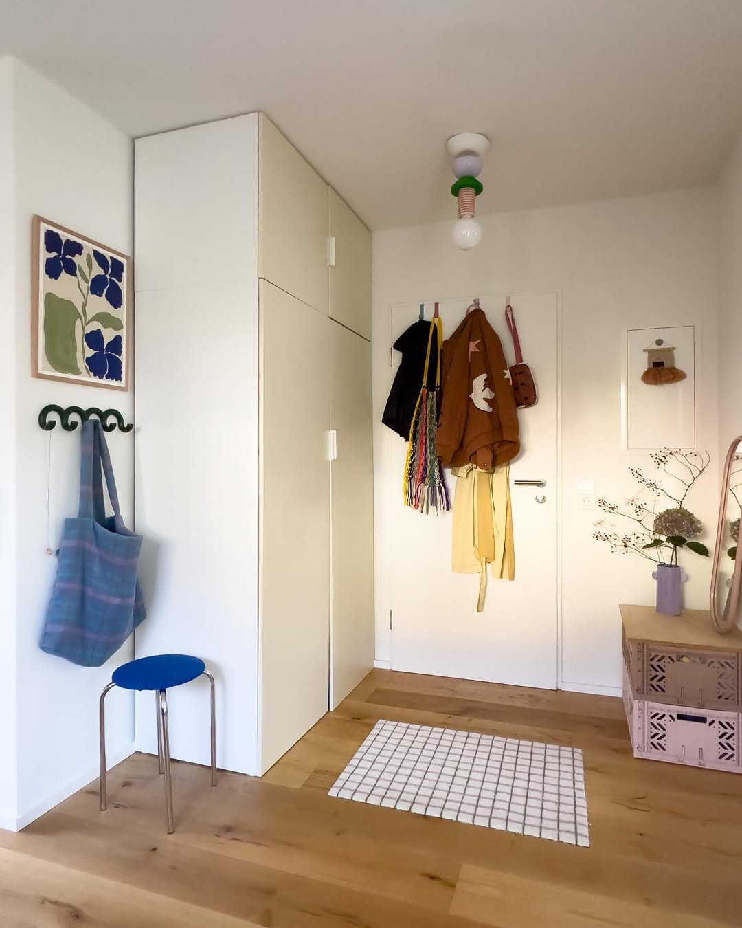 A tidy entryway with hanging coats and bags, a small stool, storage cabinets, a mat, decorative boxes, and wall art in a bright, modern room—perfect for inspiring small space decor and apartment organization.