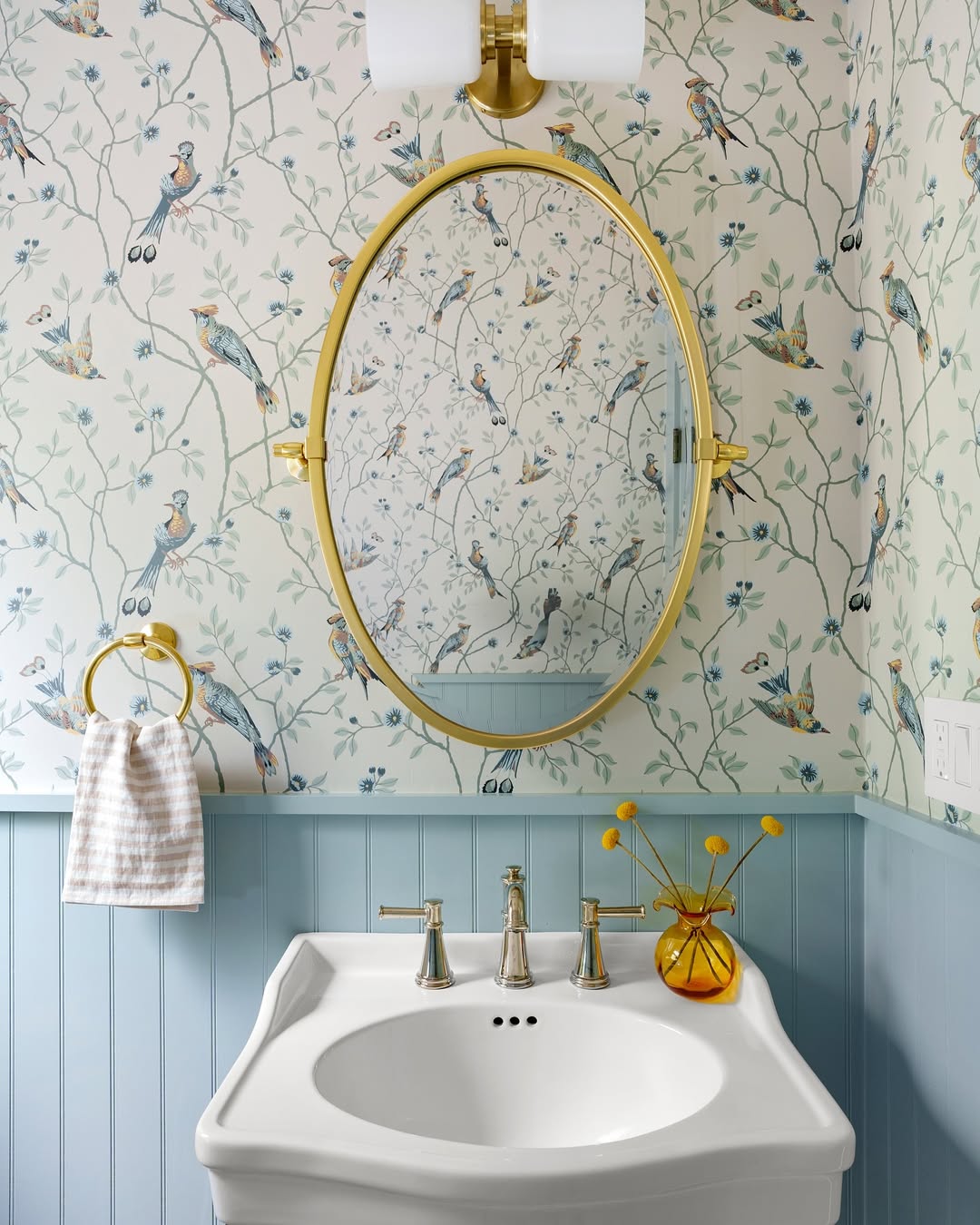A bathroom with bird-patterned wallpaper, a gold-framed oval mirror, a white sink, silver faucets, and a yellow vase with flowers on the counter—perfect inspiration for stylish powder rooms and wow-factor decor.