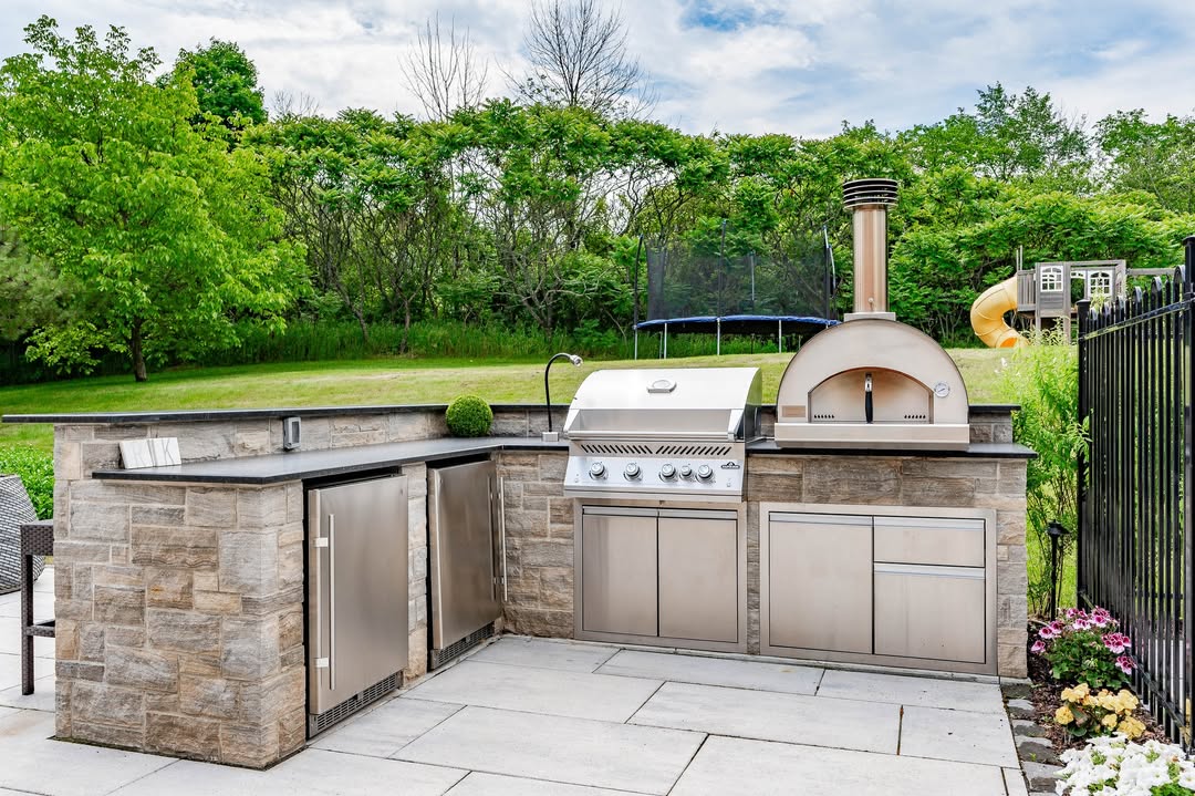 Rustic outdoor kitchen featuring a stainless steel grill, pizza oven, sink, and stone cabinetry on a patio, with green lawn and trees in the background—perfect for those seeking rustic kitchen ideas.