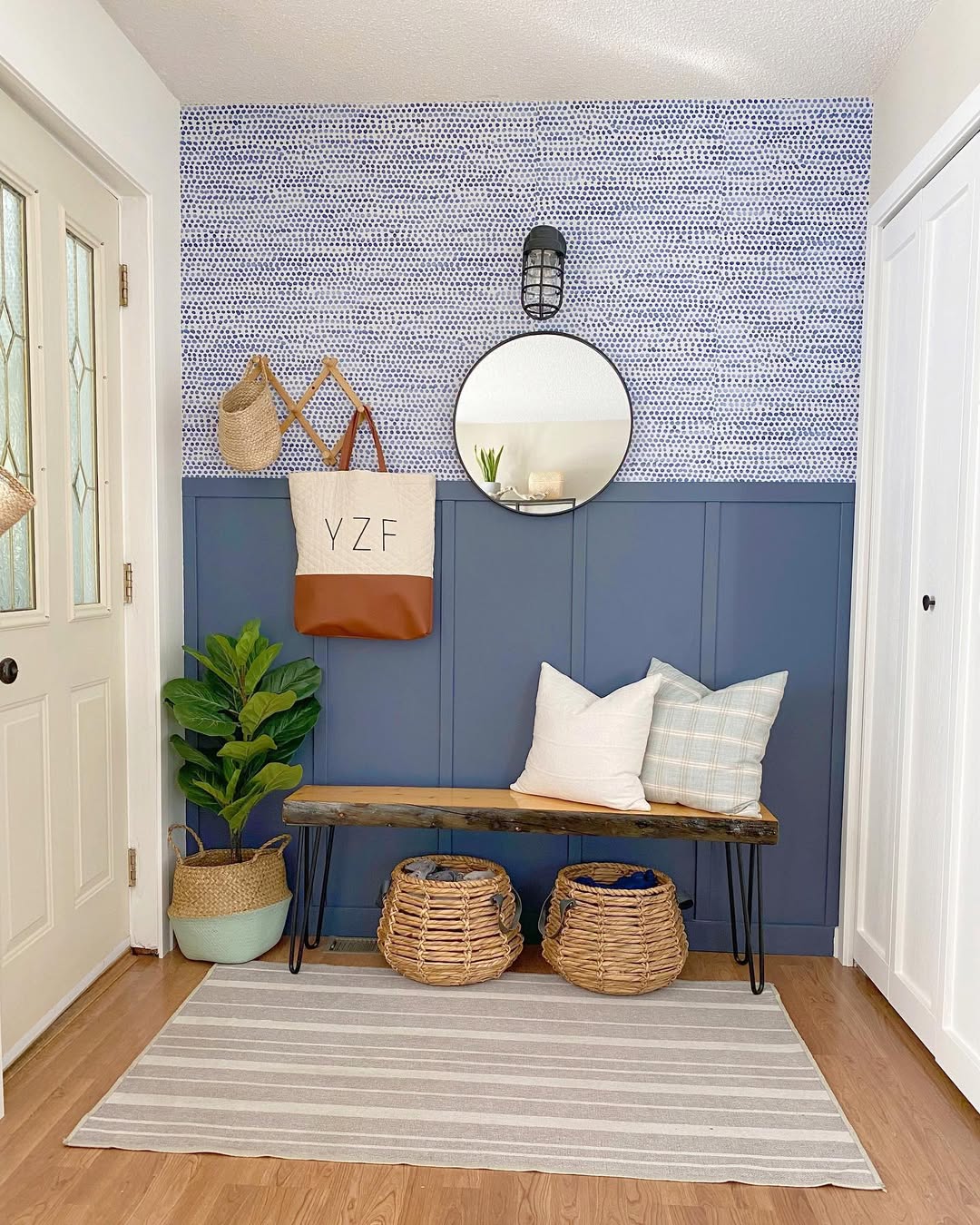 A tidy small entryway features a wooden bench, two wicker baskets, pillows, a potted plant, a striped rug, a round mirror, and blue and white walls.