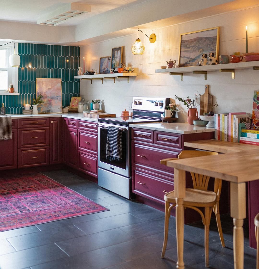 A kitchen with dark red cabinets and a teal tile backsplash showcases an eclectic touch, almost like a purple kitchen dream. Wooden shelves hold decor above a cozy table with a chair. A rug adorns the dark floor, while candles and lights provide warm illumination.
