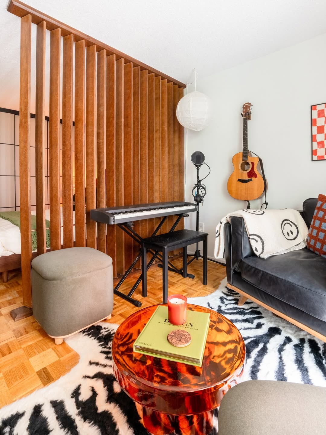A modern living space showcases clever small space decor—a keyboard on a stand, a sofa, mounted guitar, red coffee table with candle and book, and zebra print rug—all smart touches for stylish apartment design. Wood slats partially divide the room.