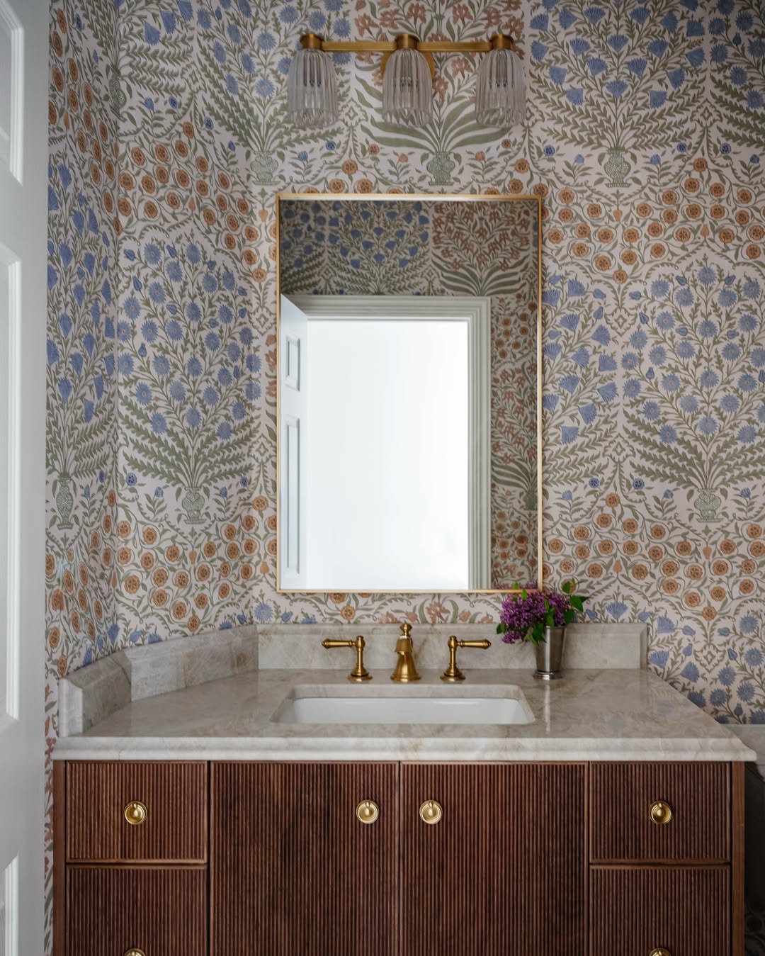 A bathroom vanity with a marble countertop, brass fixtures, floral wallpaper, a rectangular mirror, wall-mounted light fixture, and a small vase with purple flowers—perfect inspiration for stylish powder rooms.
