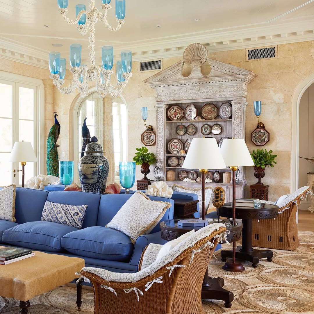 A bright living room with blue sofas, wicker chairs, blue glass accents, a large decorative shelf with plates, peacock statues, and beige stone walls—perfect inspiration for island living room ideas.