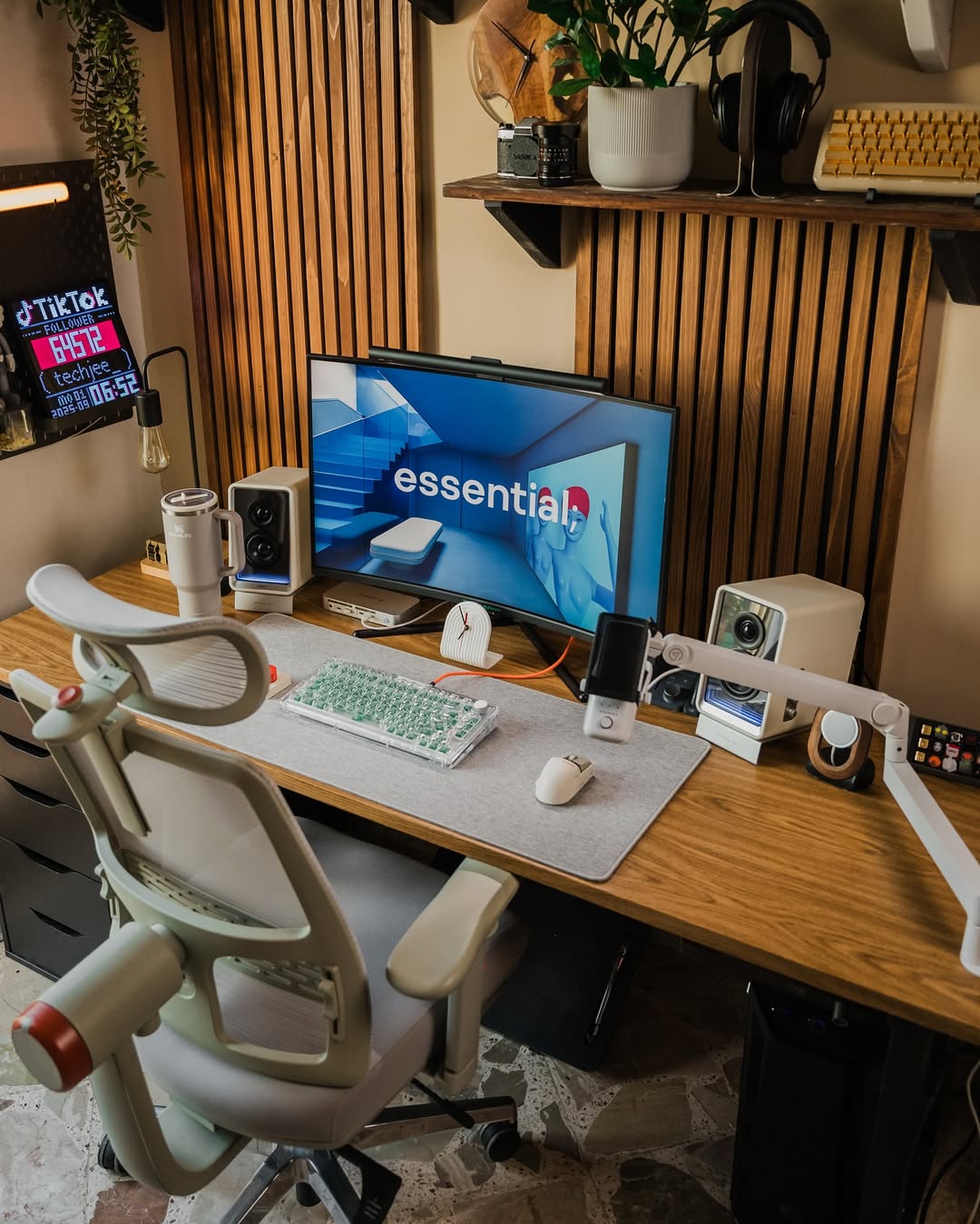 A modern home office setup with a large monitor displaying “essential,” ergonomic chair, keyboard, mouse, speakers, and various tech gadgets on a wooden desk with decorative shelves above—perfect for inspiring desk organization.