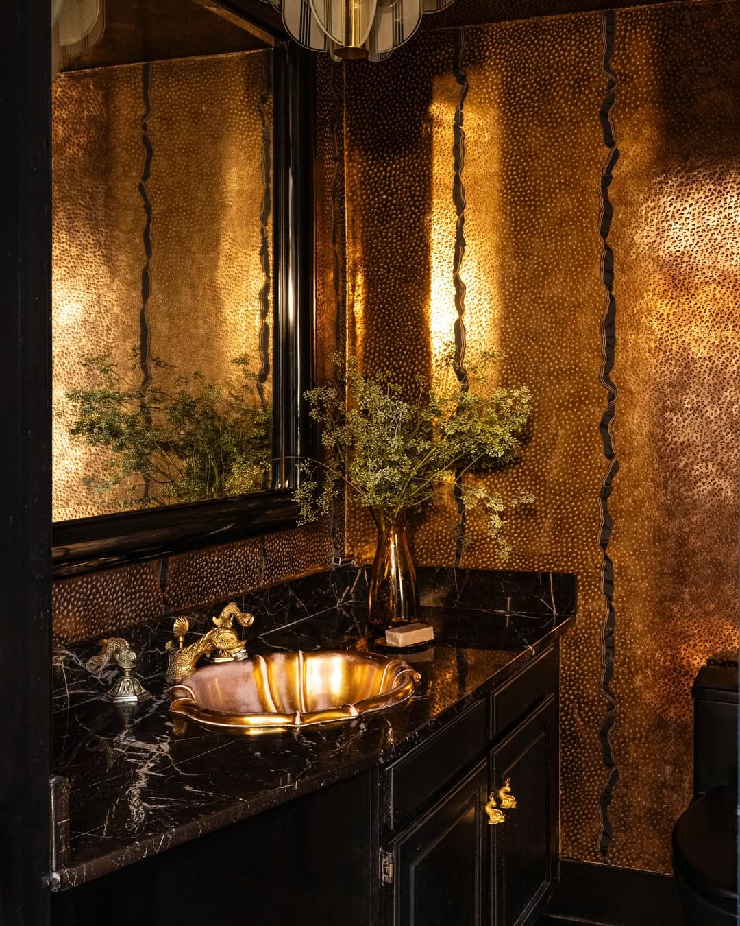 A bathroom with textured metallic gold walls, a black marble countertop, a gold sink, and a vase with greenery beside a large framed mirror—perfect for those seeking stylish powder rooms with undeniable wow-factor.