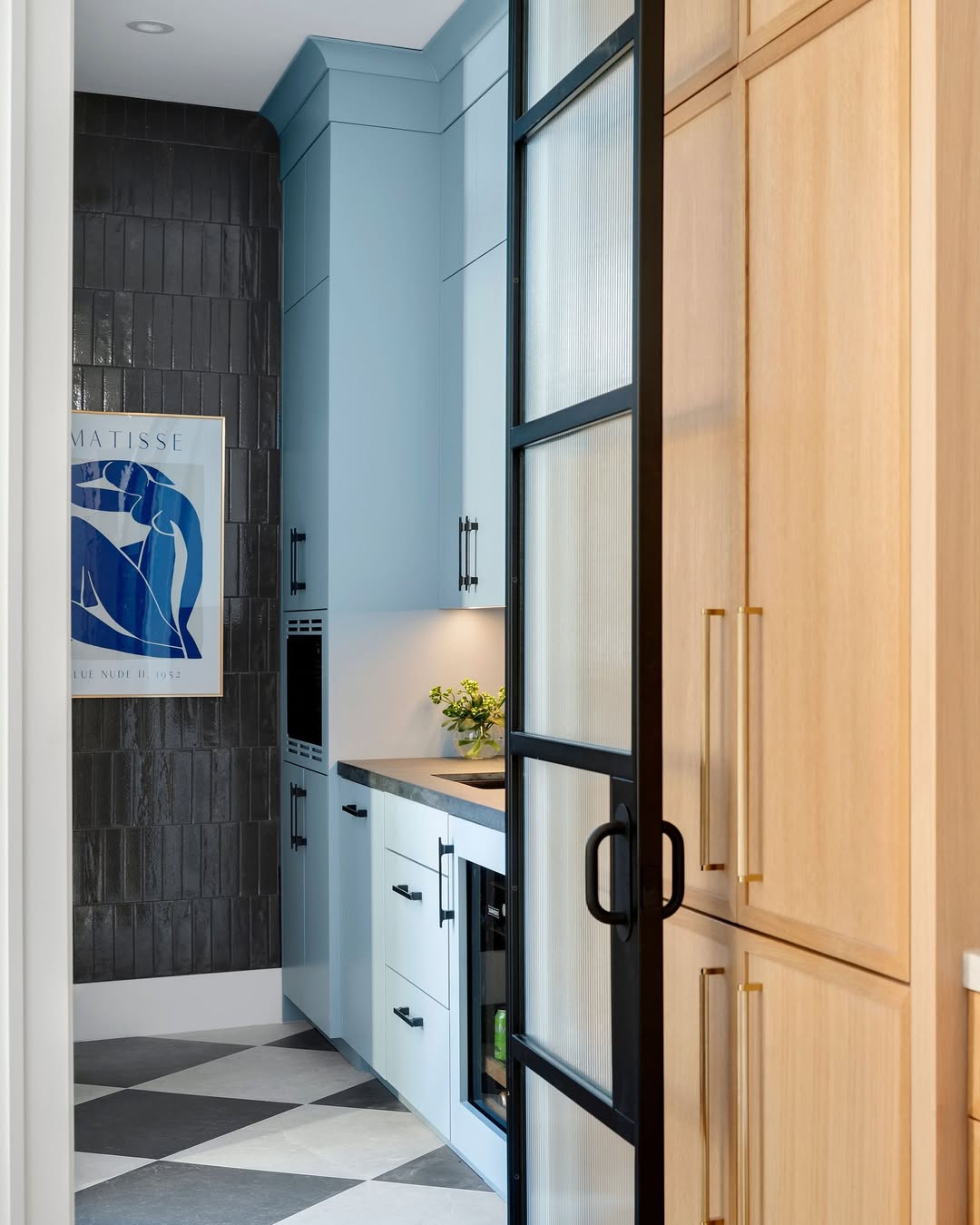 Modern galley kitchen with blue cabinets, black hardware, light wood pantry design, black-framed glass door, dark tiled wall, checkered floor, and a blue Matisse art print.
