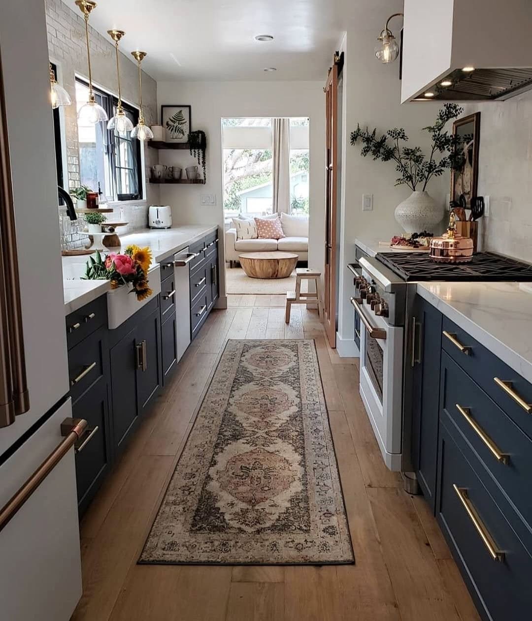 A modern galley kitchen with dark blue cabinets, brass hardware, white countertops, and hanging lights showcases smart small kitchen design. A patterned rug lines the floor, leading to a bright breakfast nook at the far end.