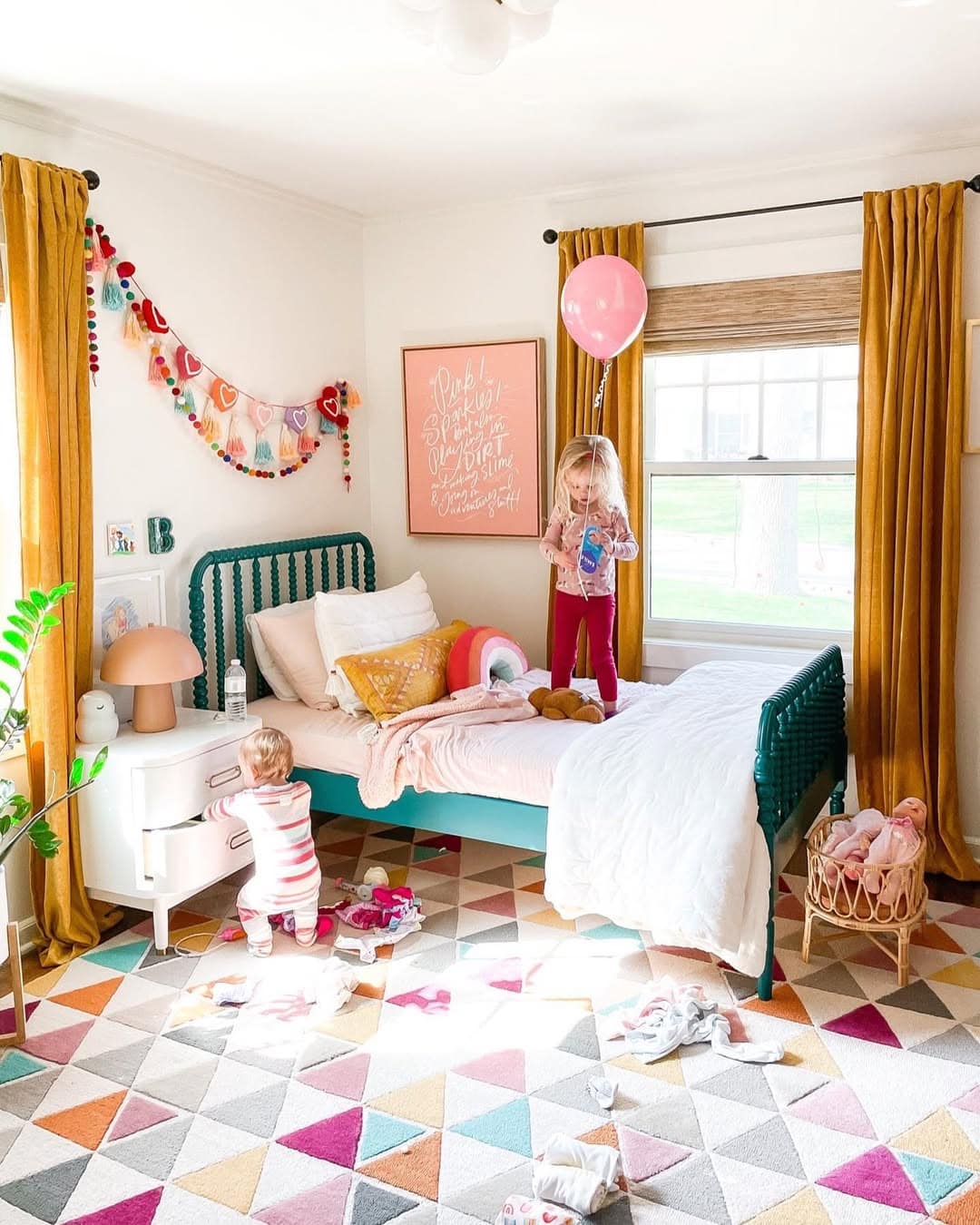Two children joyfully play in a cute toddler room, featuring a geometric-patterned rug, orange curtains, and a green bed. One child holds a balloon on the bed while the other engages with toys on the floor, offering delightful bedroom ideas for a vibrant space.