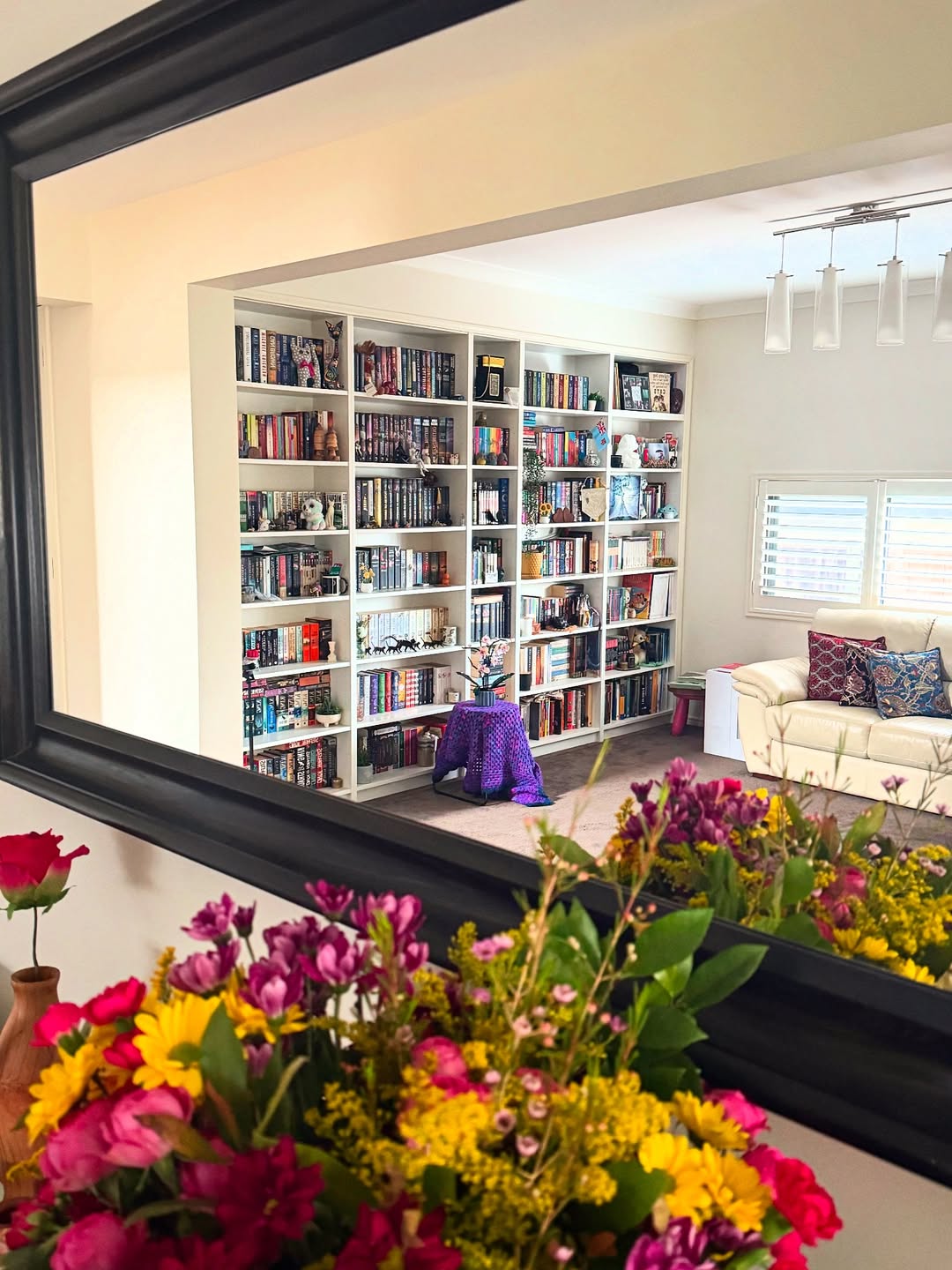 Reflection in a mirror shows a living room with standout bookshelf decor, a large shelf filled with books, a white couch, and a small round table; a bouquet of flowers is in the foreground.