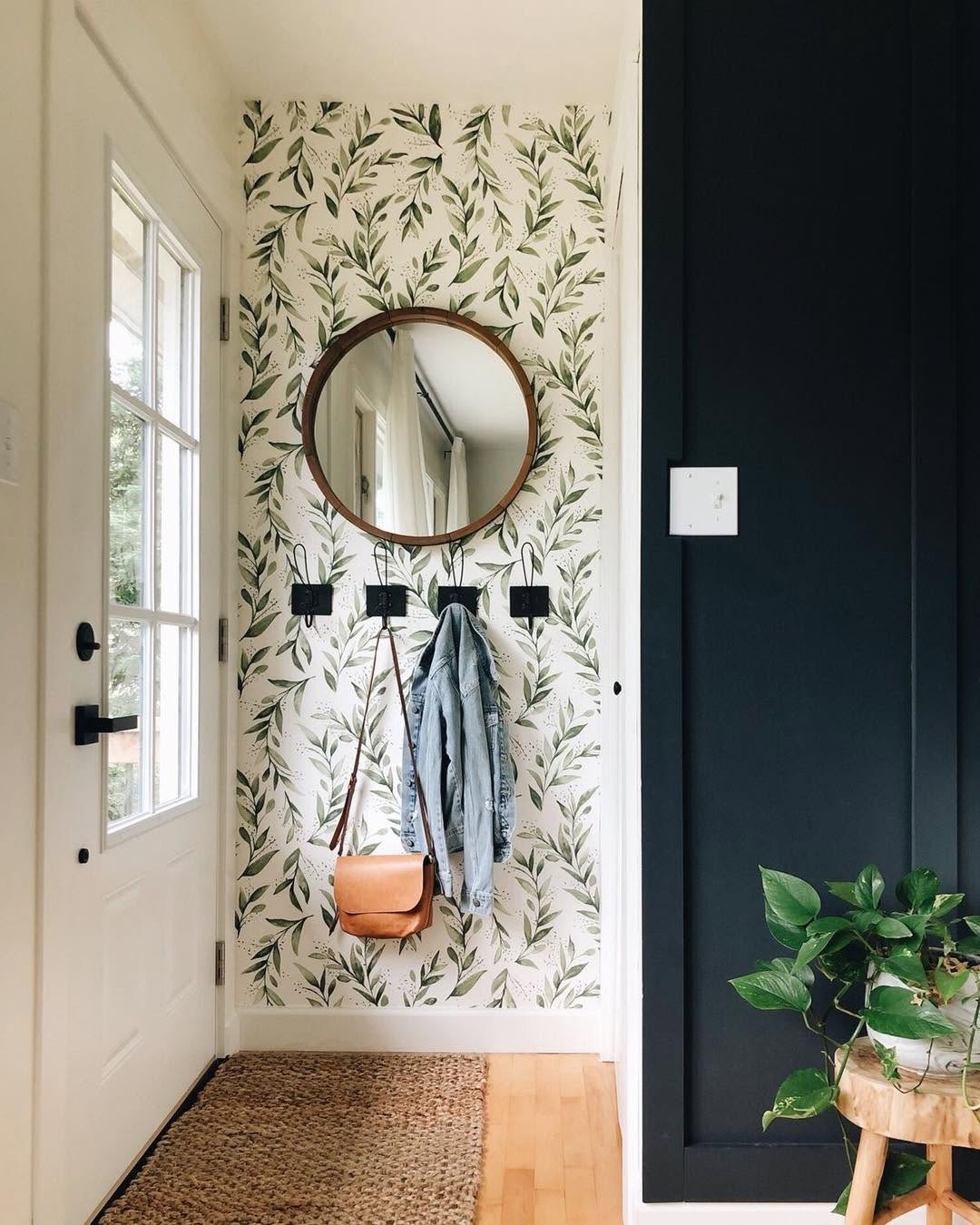 A small entryway features a leafy wallpaper accent wall, round mirror, and coat hooks holding a jacket and bag. A plant sits on a wooden stool by the dark painted wall, making this small entryway feel organized and welcoming.