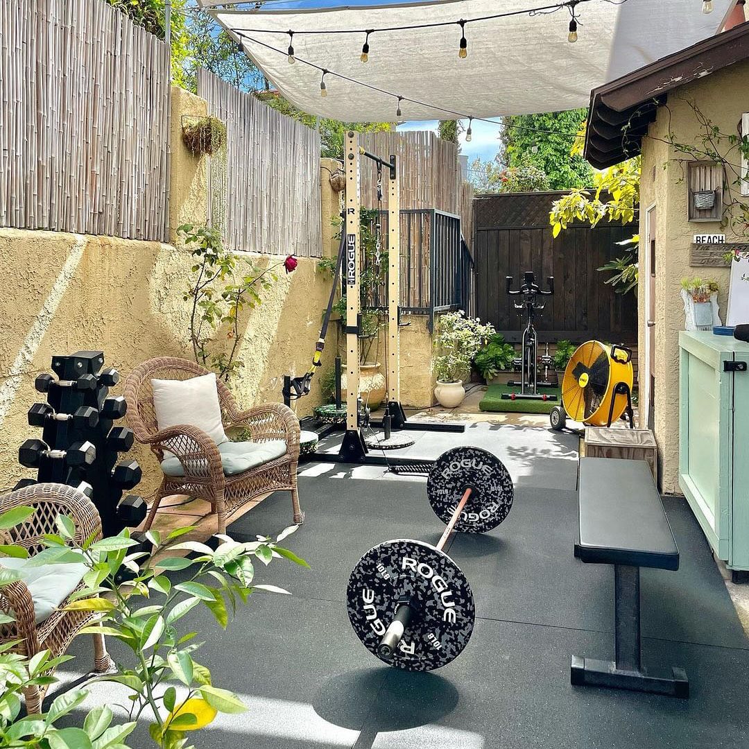 Outdoor mini gym at home featuring a barbell, bench, dumbbells, stationary bike, and rattan chairs under a sunshade, all surrounded by bamboo fencing and lush greenery.