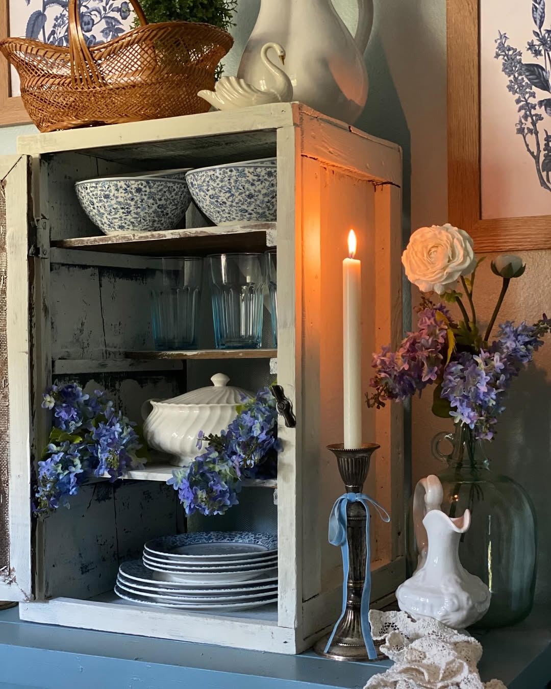 A vintage white cabinet displays blue floral dishes, glassware, and a teapot. A lit candle, blue flowers, and a white figurine rest nearby—perfect inspiration for charming Vintage Farmhouse Decor.