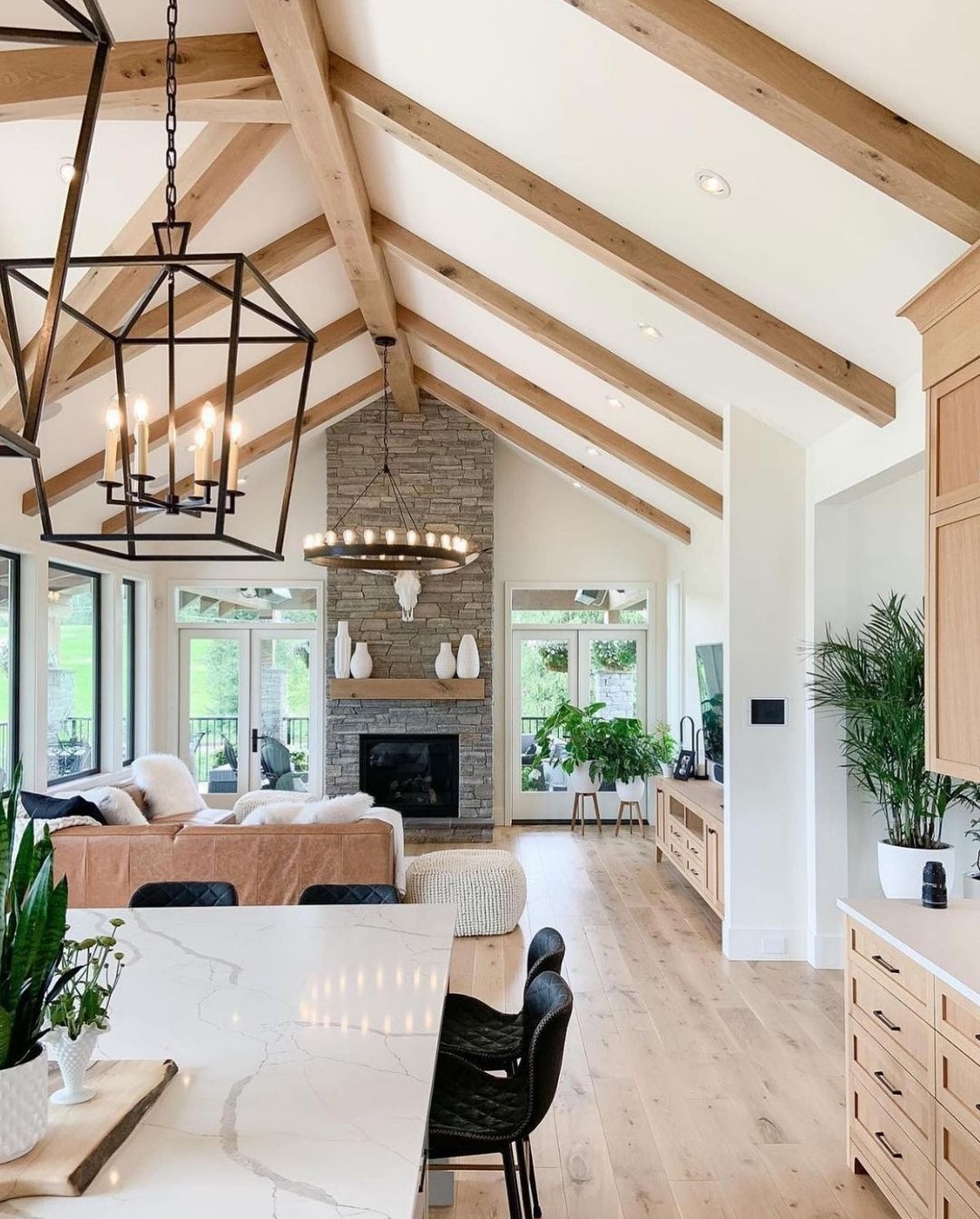 Open-concept living room and kitchen with wood beams, large windows, stone fireplace, modern lighting, light wood floors, and indoor plants—perfect high ceiling living room ideas for a brighter, bigger home feel.