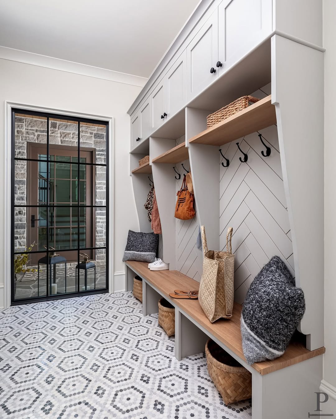 A genius mudroom with built-in cubbies, wooden benches, storage baskets, wall hooks, and a patterned tile floor; glass door opens to a stone-walled exterior—perfect entryway inspiration for smart organization.