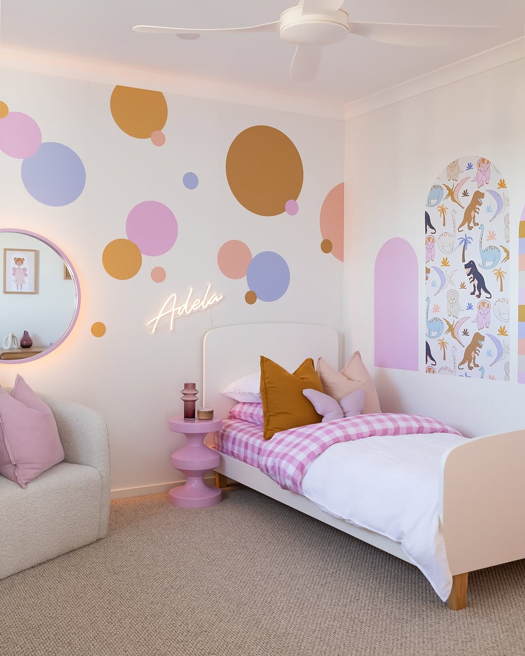 A toddler girl bedroom with a white bed adorned in pink and orange bedding and colorful circle wall decals. A dinosaur-themed arch accents one wall, while a neon sign spells