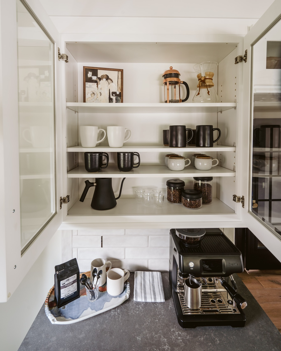 Open cabinet with shelves holding mugs, a French press, glass jars, and a coffee grinder above a countertop organization setup with an espresso machine, coffee beans, and utensils—perfect for inspiring Kitchen coffee bar ideas.