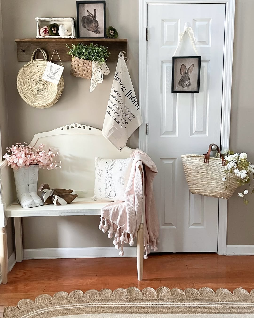 A white bench with pink and white decor sits in a small entryway against a beige wall; baskets, artwork, and flowers hang on nearby hooks and a white door.