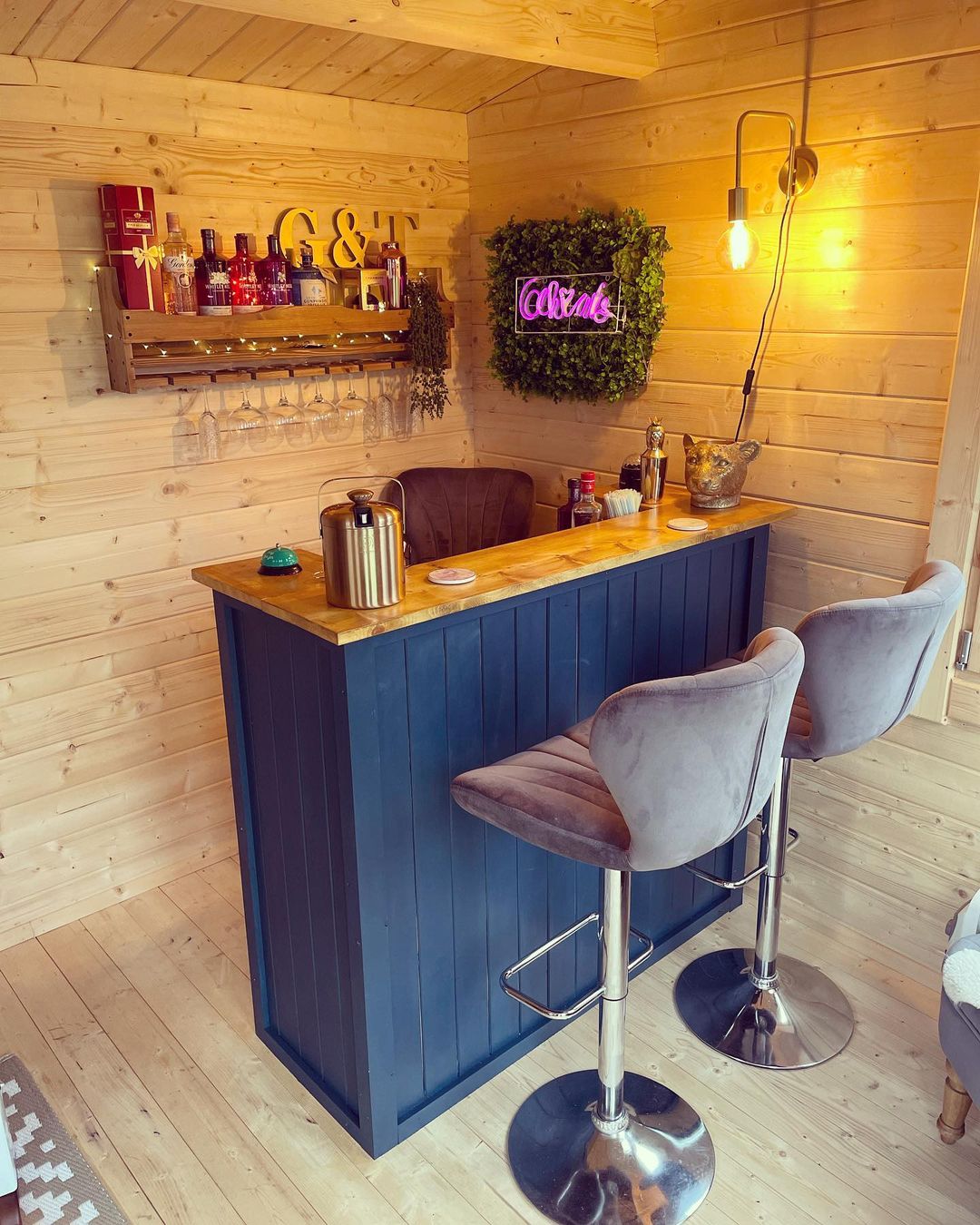A small home bar with a wooden counter, two grey bar stools, wall-mounted shelves with bottles, glassware, a neon
