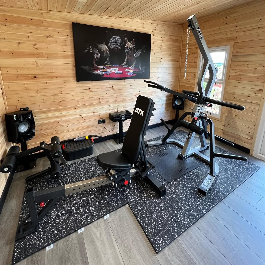 A mini gym at home featuring an adjustable ATX weight bench, leg extension machine, and weightlifting station on a rubber mat in a wooden-paneled room, complete with speakers and dog-themed artwork on the wall.