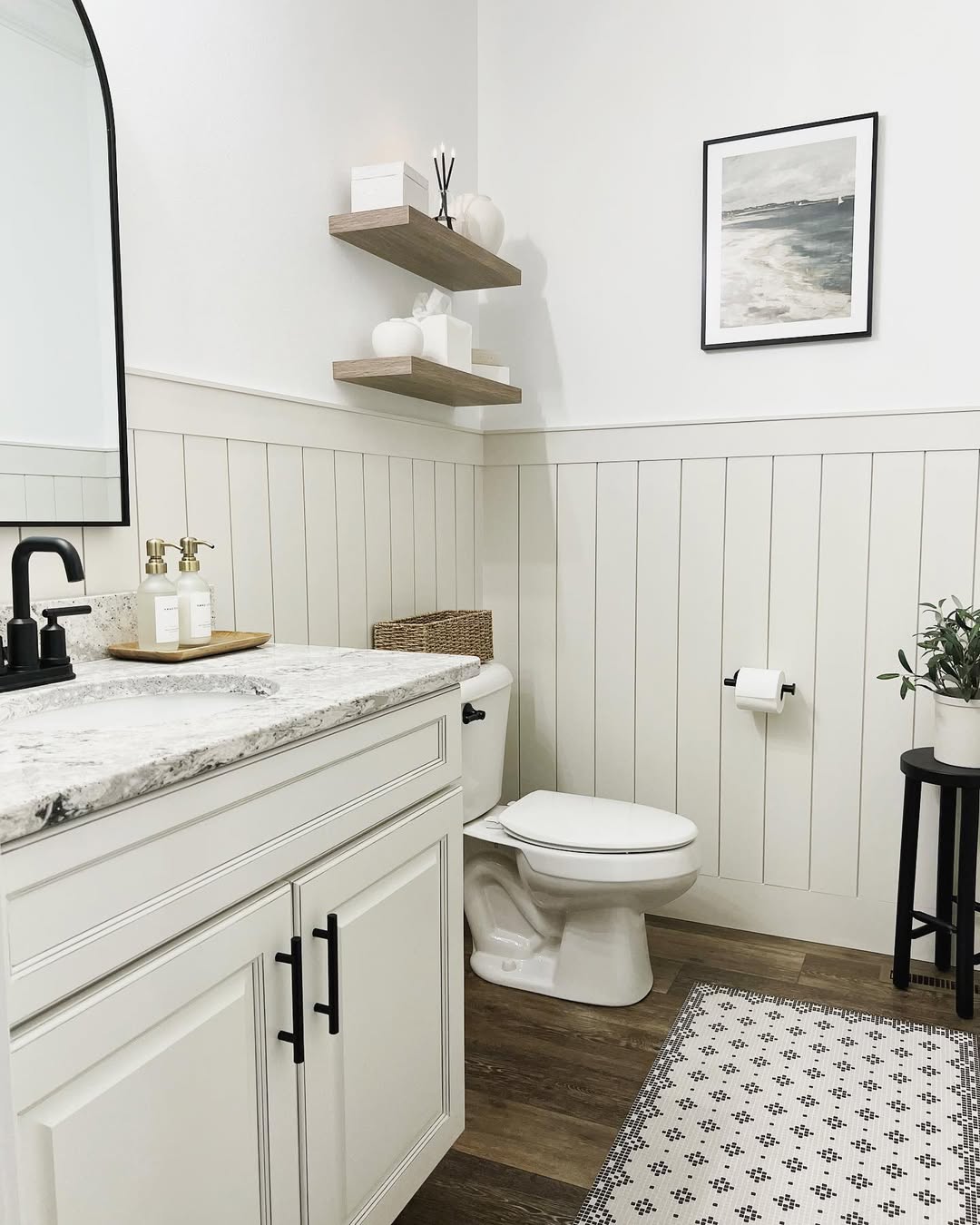Bathroom with white shiplap wall paneling, a marble countertop, black fixtures, floating shelves with decor, a plant on a small table, and a framed ocean print on the wall.