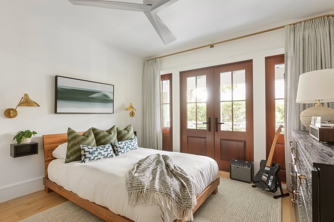 A modern boys bedroom with a wooden bed, green and patterned pillows, a blanket, wall art, French doors, curtains, a dresser offering storage solutions, a lamp, a guitar, and an amplifier on the floor.