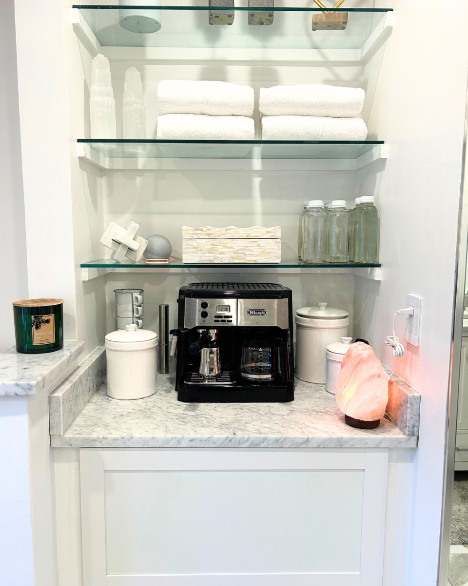 A coffee station with an espresso machine, cups, jars, a candle, and a salt lamp sits on a marble counter. Thoughtful countertop organization and stylish coffee bar decor add charm to the shelves holding towels and decorative items.
