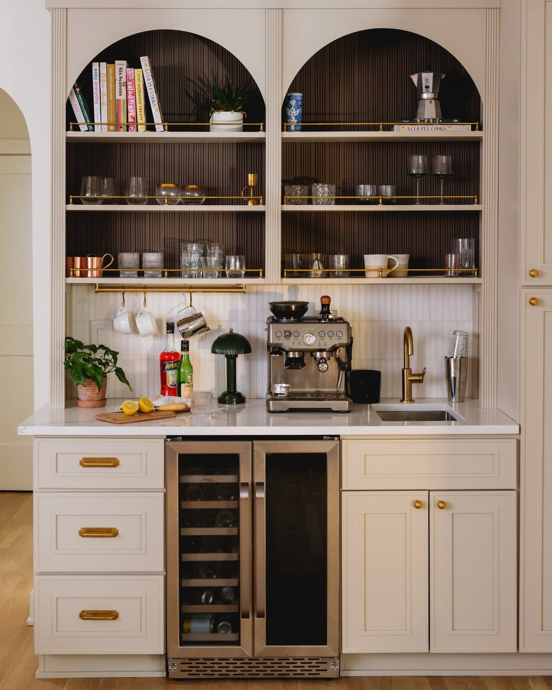 A home coffee bar with built-in shelves for glassware and books, a countertop espresso machine to tame countertop chaos, a sink, plants, and a dual-zone wine fridge below—perfect inspiration for your kitchen coffee bar ideas.