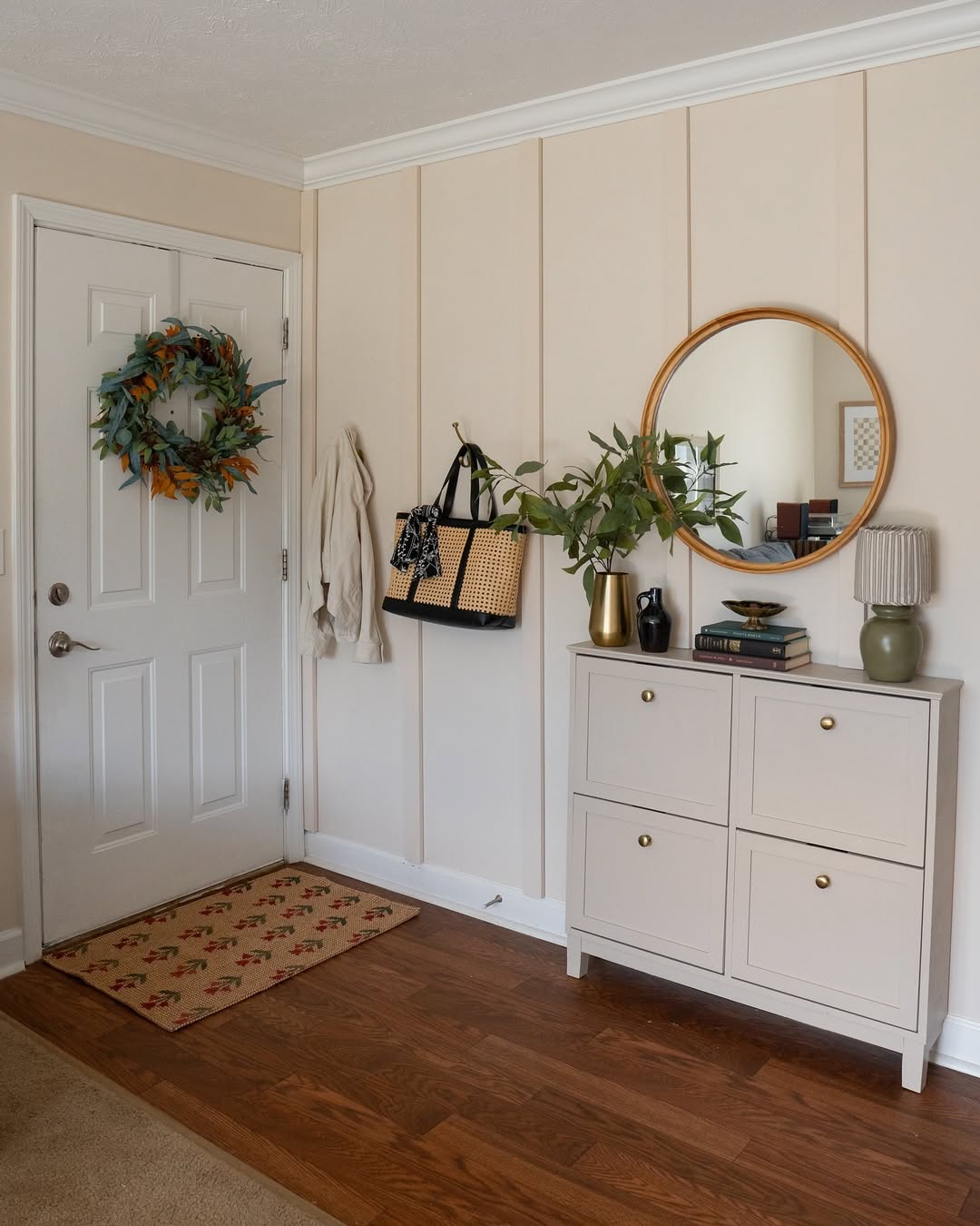 This small entryway features a white door with a green wreath, wall hooks holding bags and a coat, a shoe cabinet adorned with decor, and a round mirror for added charm.