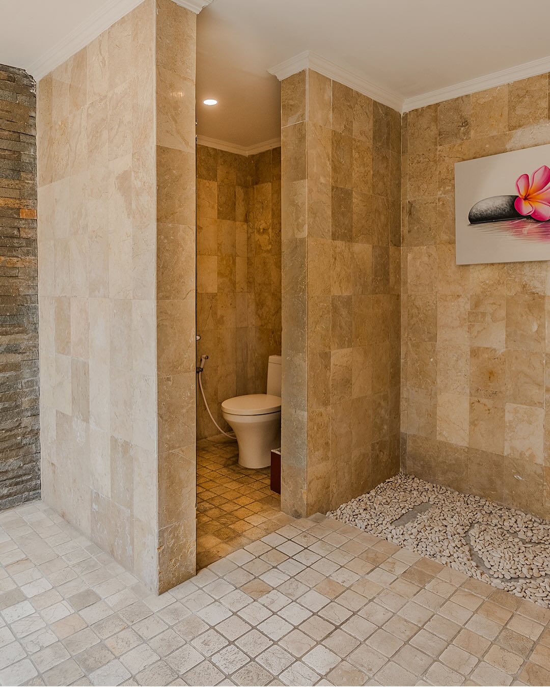 A modern small bathroom with beige stone tiles features a no door shower area lined with pebbles and a toilet in a partially enclosed section; a framed photo decorates the wall.