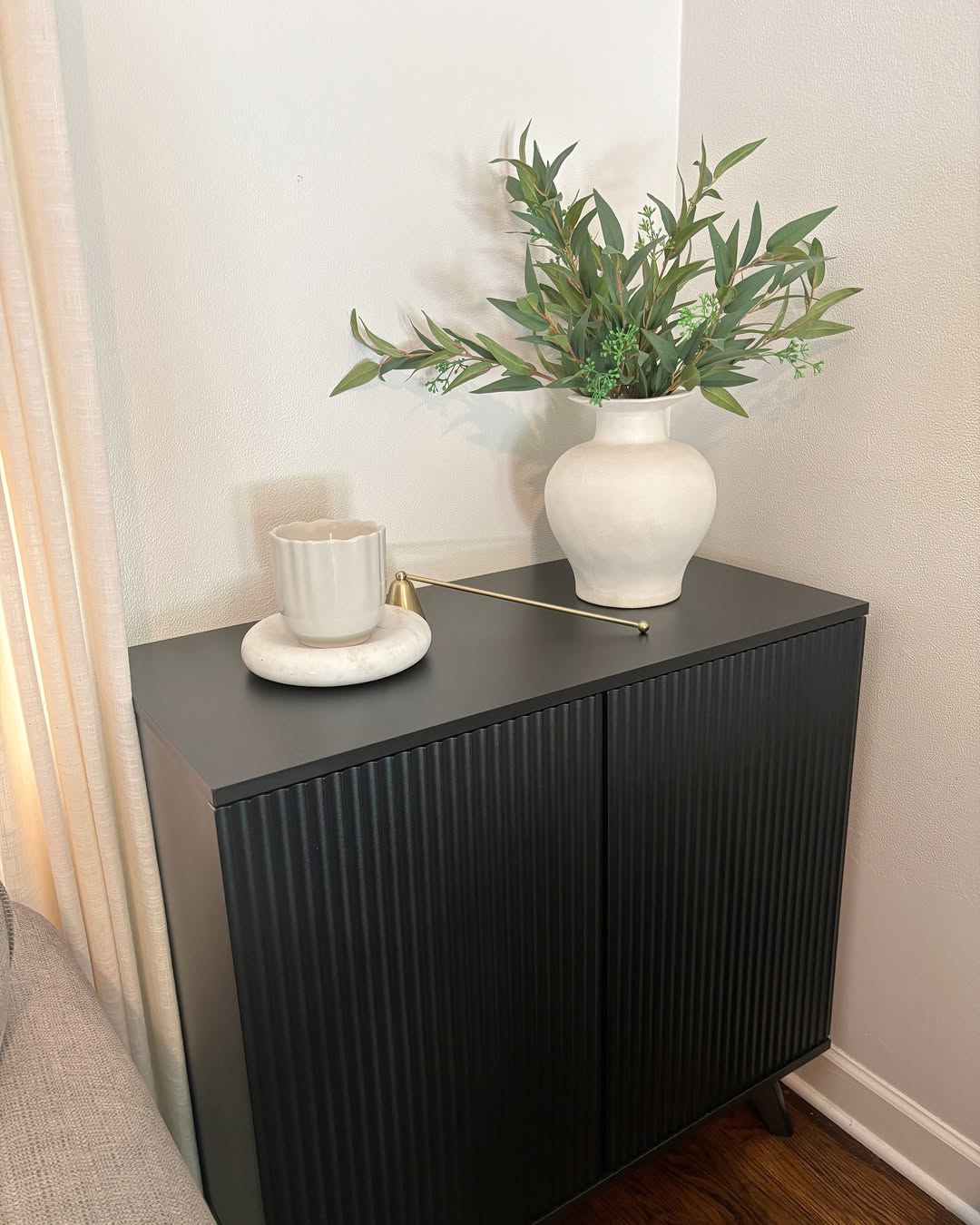 A black cabinet styled with sideboard decor—a white vase of greenery, a round tray, mug, and gold candle snuffer—sits in the corner of a white-walled room, offering simple and elegant decor ideas.