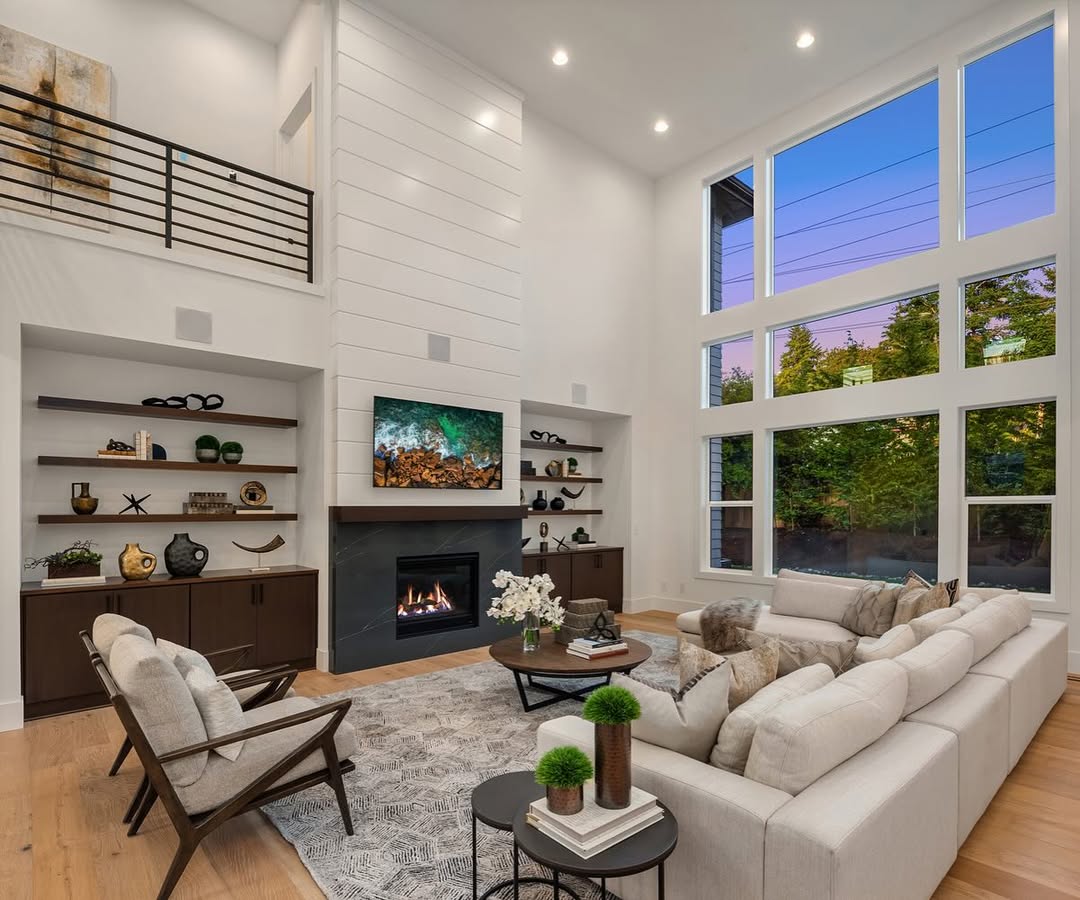 Modern living room with a high ceiling, large windows, a sectional sofa, fireplace under a mounted TV, built-in shelves, and neutral-colored decor—a perfect example of high ceiling living room ideas for a bigger home.
