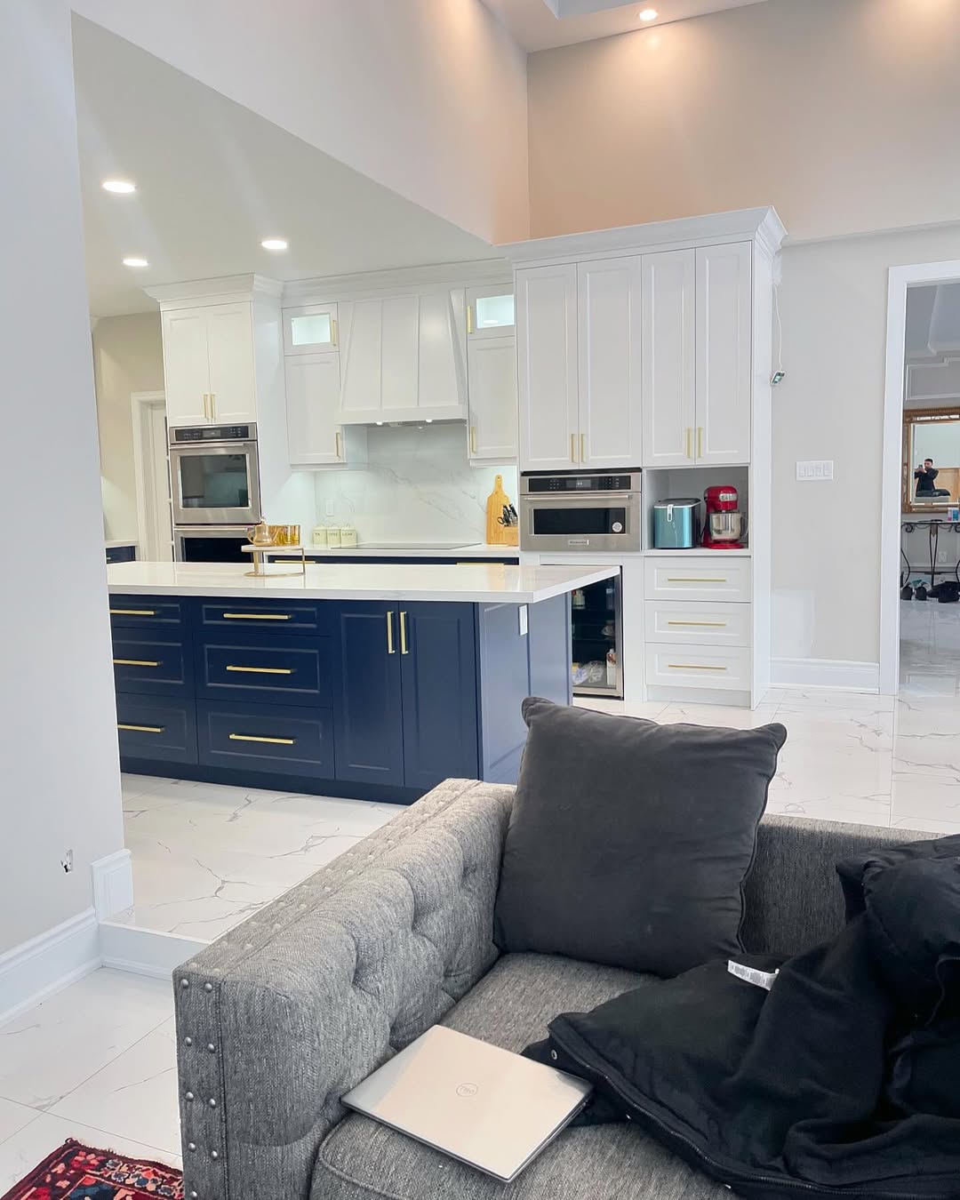 Modern kitchen with white upper cabinets, navy lower cabinets, gold handles, built-in appliances, marble countertops, and a gray tufted sofa in the foreground with a laptop on it.