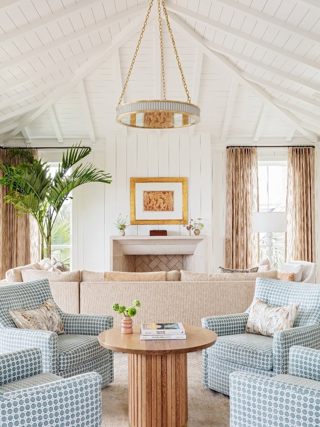 Bright living room with vaulted white ceiling, beige sofa, two patterned blue armchairs, round wooden coffee table, large plant, and fireplace with framed art above it—perfect for inspiring island living room ideas.