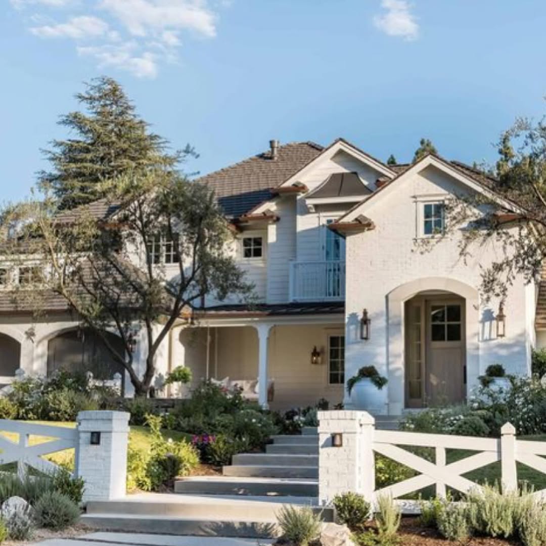 Two-story white house with a covered porch, gray roof, and landscaped front yard, viewed behind a white picket fence and classic farmhouse gate under a clear blue sky.