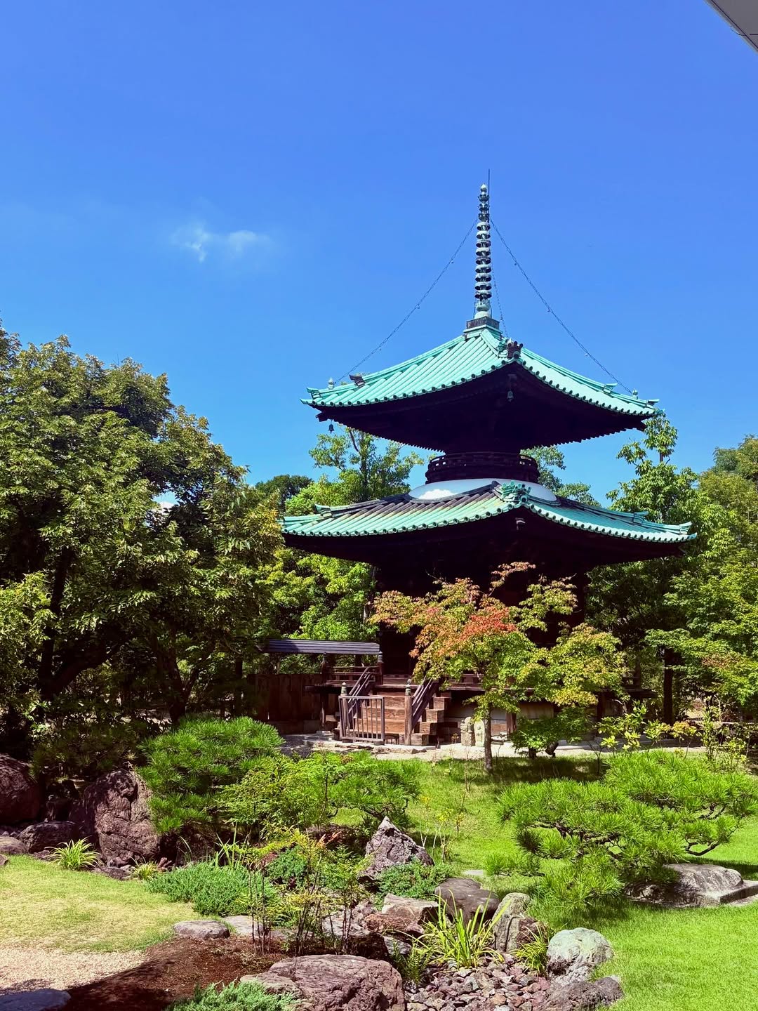 A traditional Japanese pagoda with a green roof stands amid lush trees and a landscaped Japanese garden, offering timeless garden design inspiration under a clear blue sky.