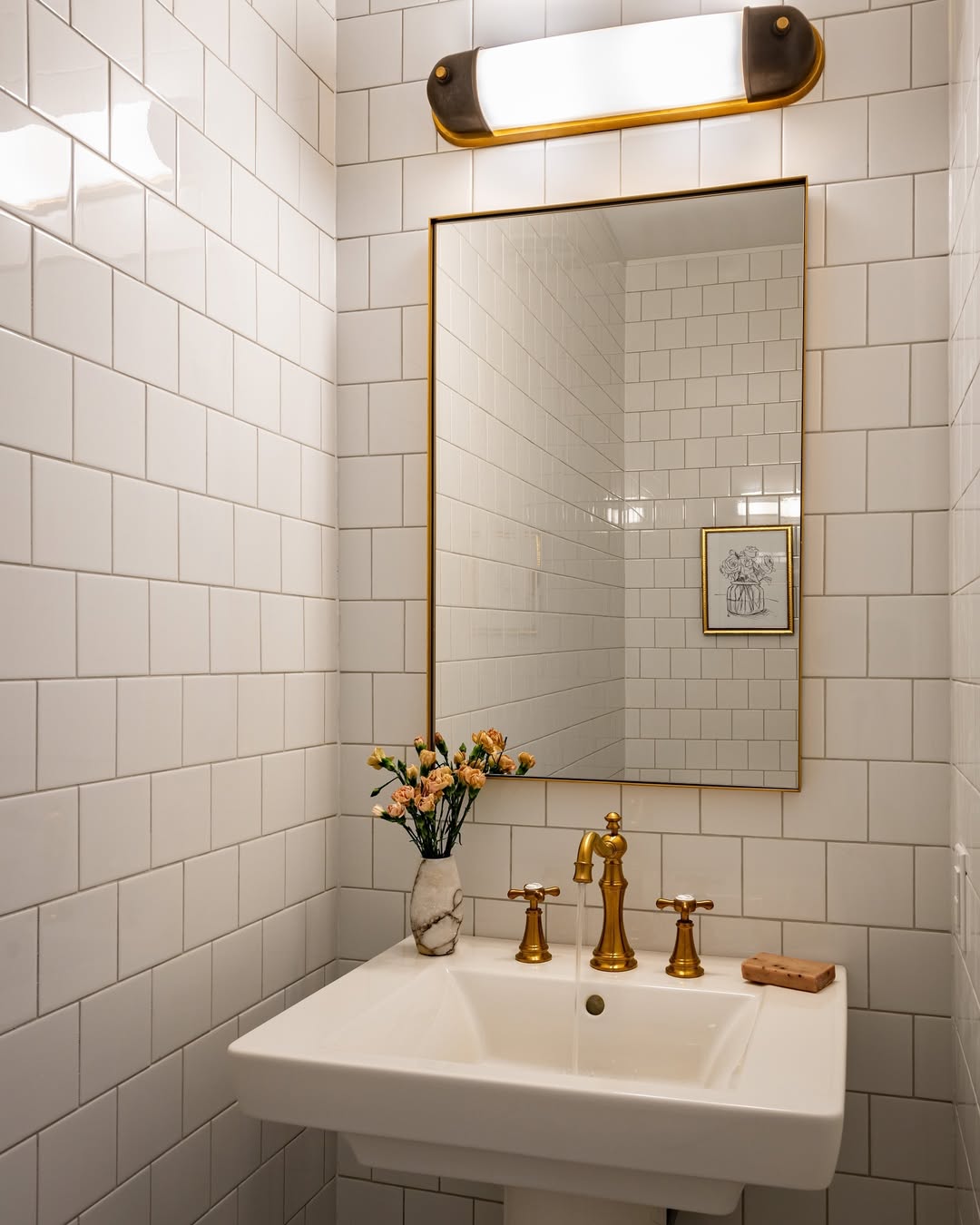 A small bathroom with white tiled walls, a rectangular mirror with a gold frame, and gold fixtures offers inspiring powder room ideas for those seeking wow-factor in stylish powder rooms. A vase of flowers and wall-mounted lighting complete the look.