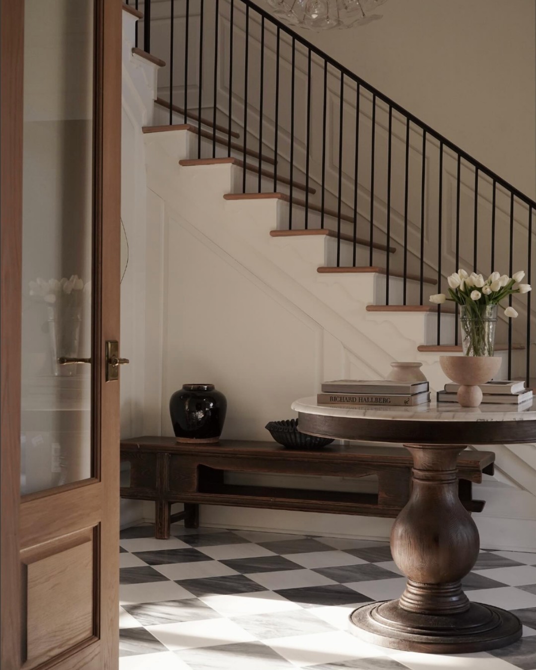 A round wooden table with books and a vase of white tulips sits on a checkered floor near a staircase with a black railing, offering fresh inspiration for entryway design in this bright, modern entryway.